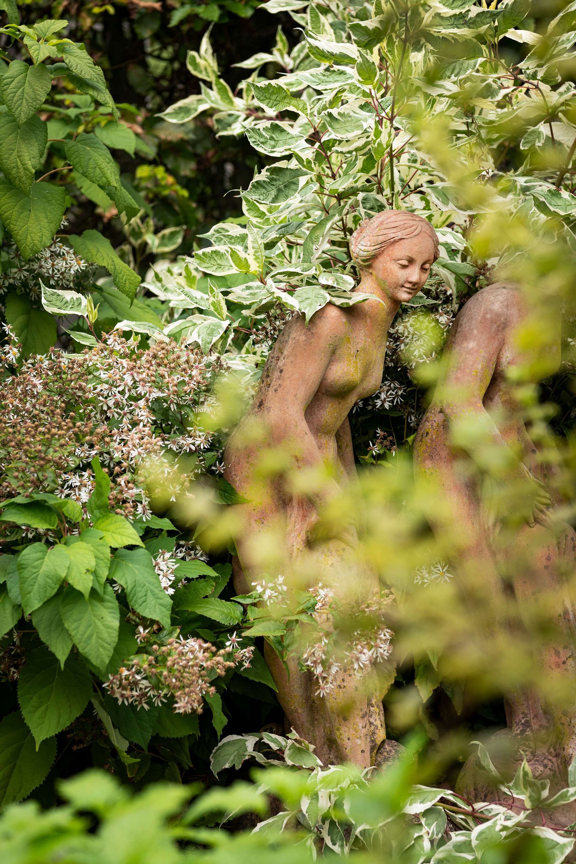Stenen sculpturen van twee figuren, gedeeltelijk verborgen door weelderig groen en gevarieerd gebladerte in een tuin.