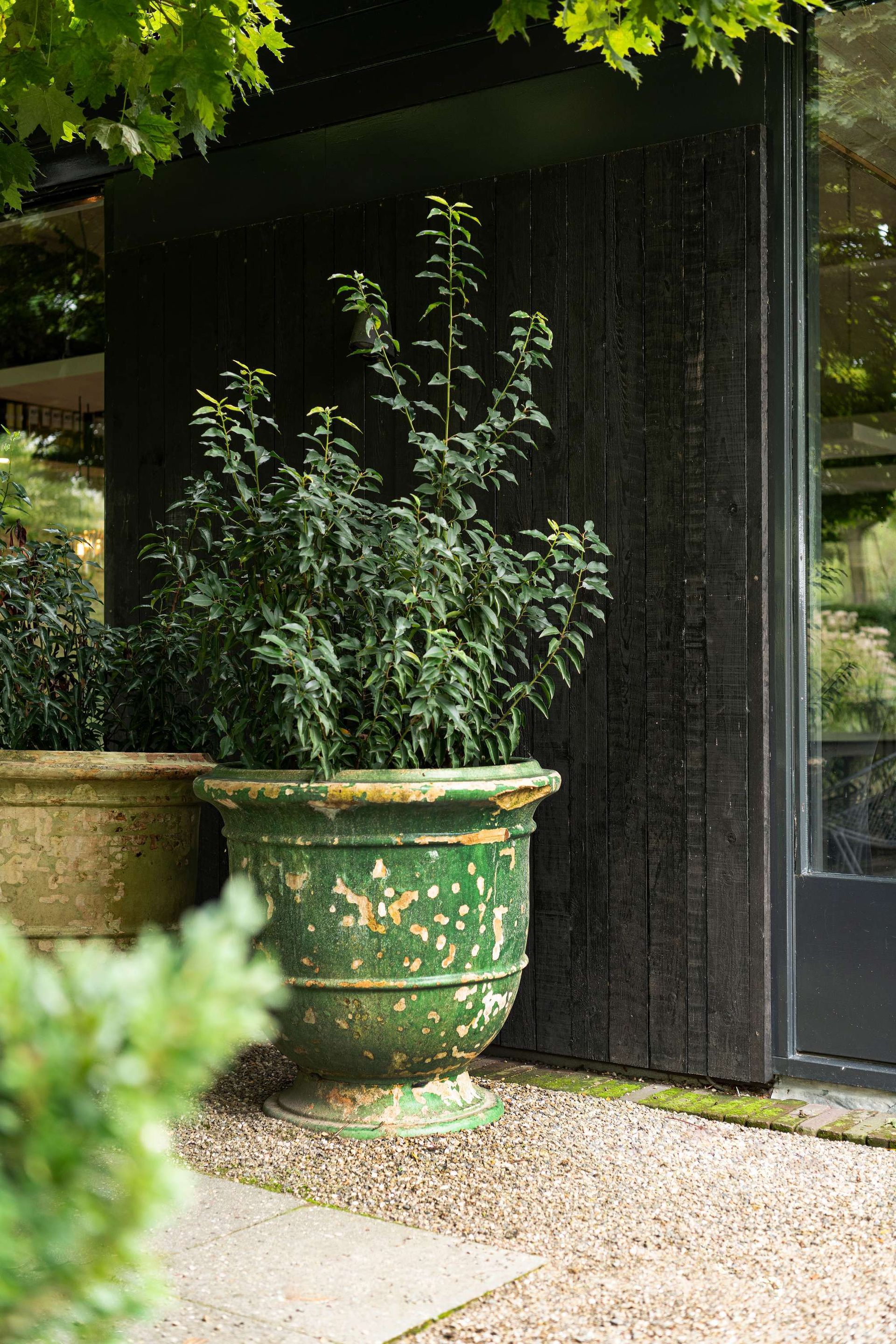 Een groene, verweerde urn met een plant staat naast een donkere muur en een raam op een grindterras.
