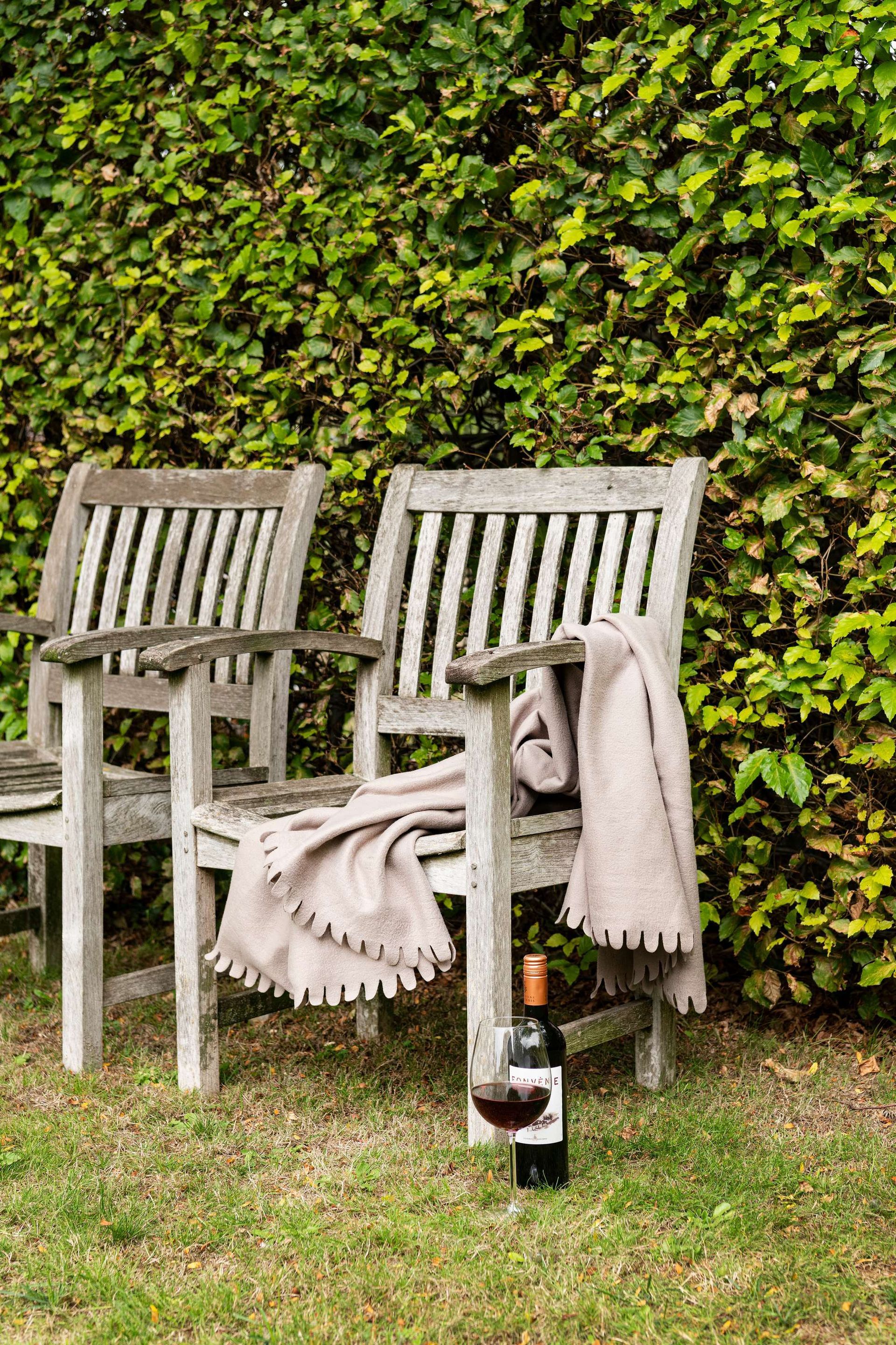 Twee verweerde houten stoelen met een deken en een fles wijn in een grasrijke tuin.