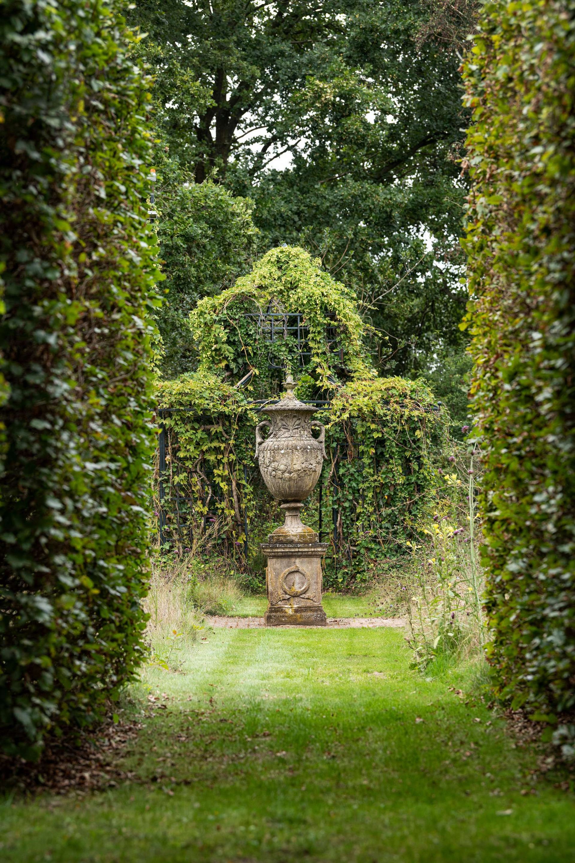 Een weelderig groen tuinpad leidt naar een sierlijke stenen urn, omgeven door hoge hagen en groen.
