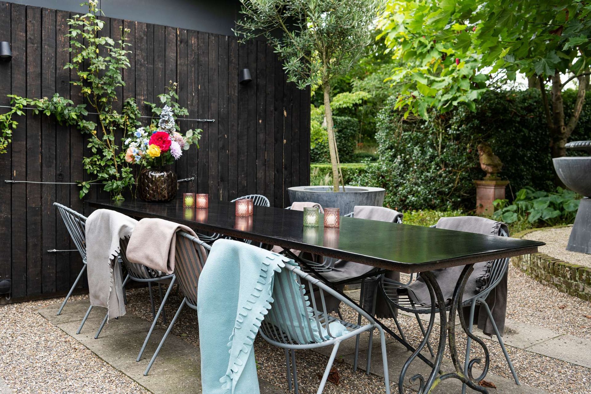 Eettafel voor buiten, gedekt voor gasten, met een donkere houten schutting en groen op de achtergrond.