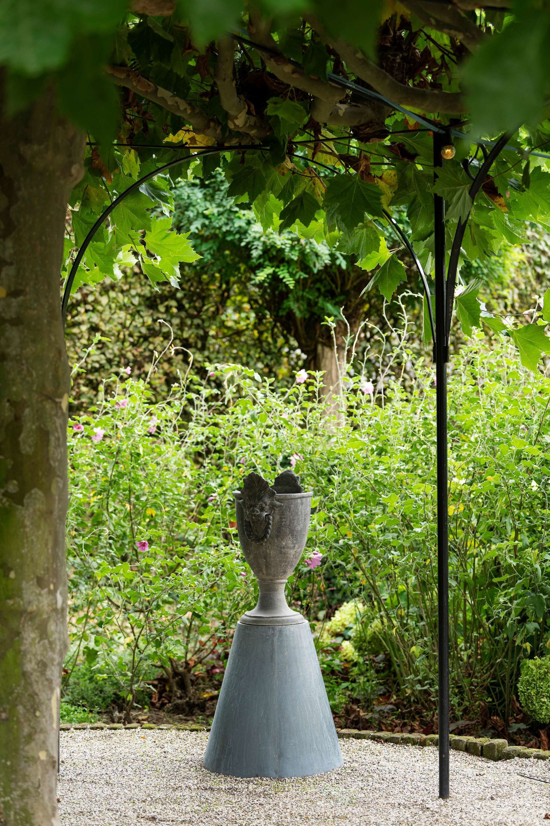 Een stenen urn op een sokkel in een tuin, onder een bladerdak van groene bladeren.