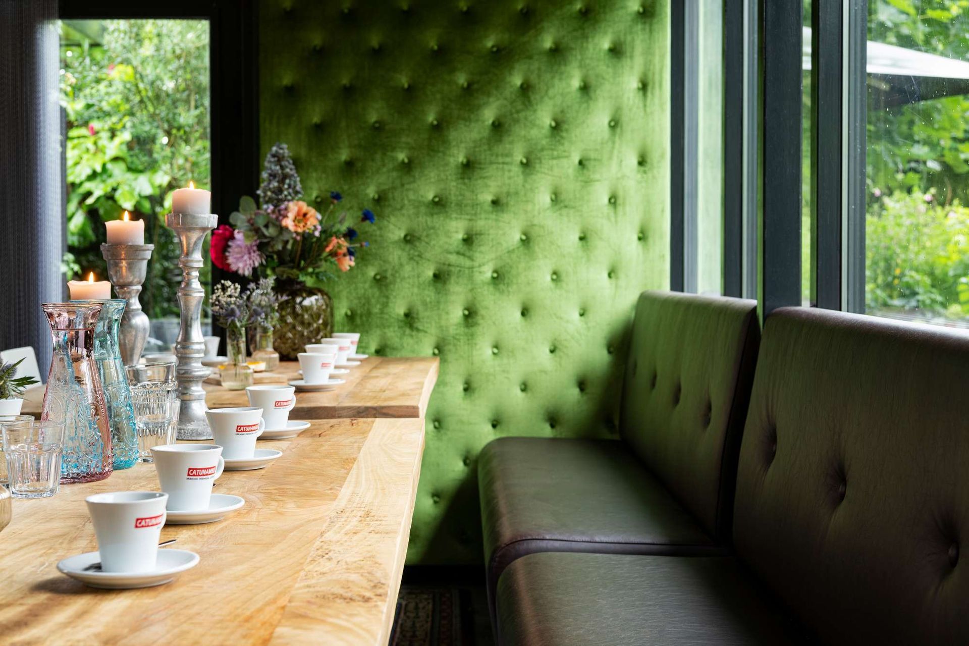 Een eettafel met koffiekopjes, kaarsen en bloemen, naast een groene gestoffeerde muur en een bankje bij een raam.