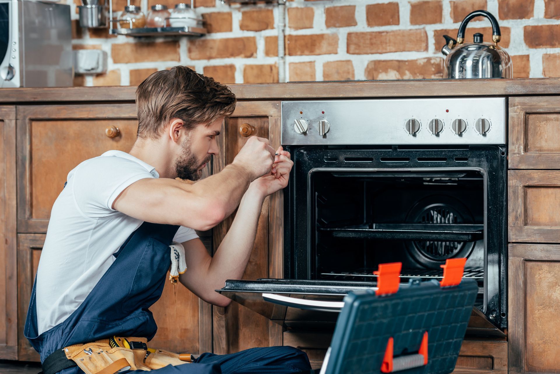 Young technician in protective workwear, performing appliance repair in a residential kitchen.