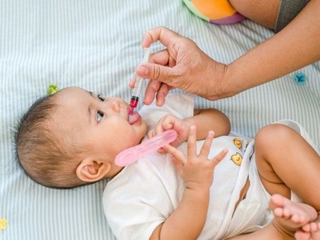 adult hand using an oral syringe to administer red liquid medication into the mouth of a baby lying on their back.