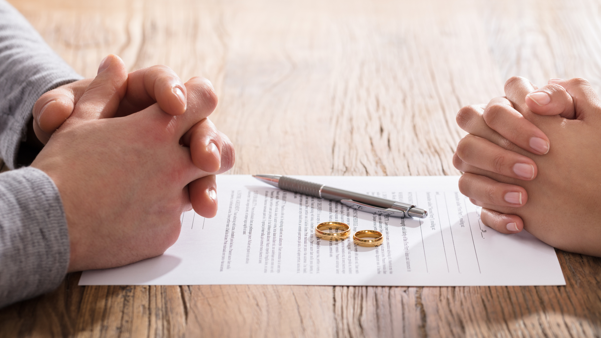 A man and a woman are sitting at a table with their hands folded over a piece of paper with wedding rings on it.