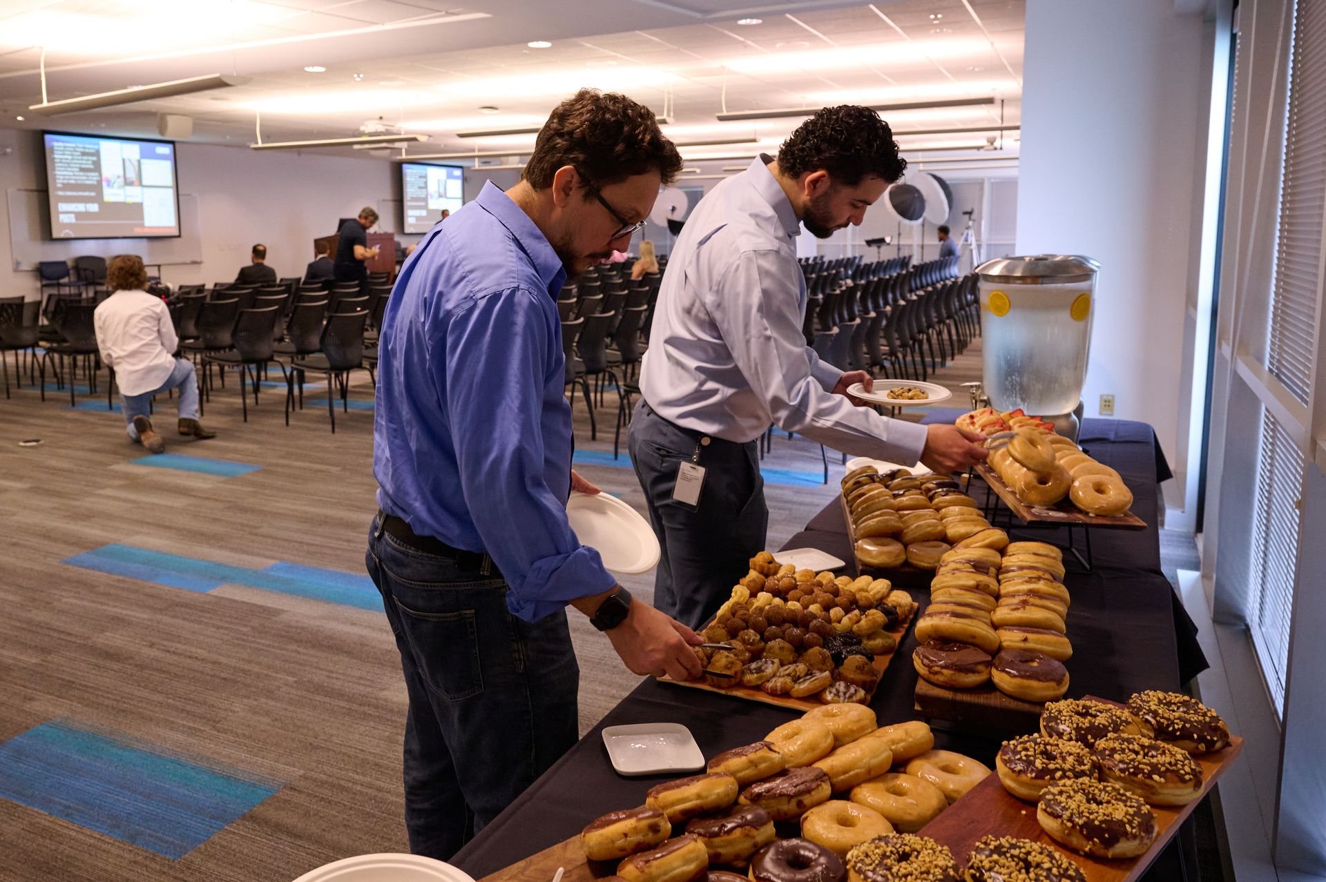 Two men are standing around a table filled with donuts.