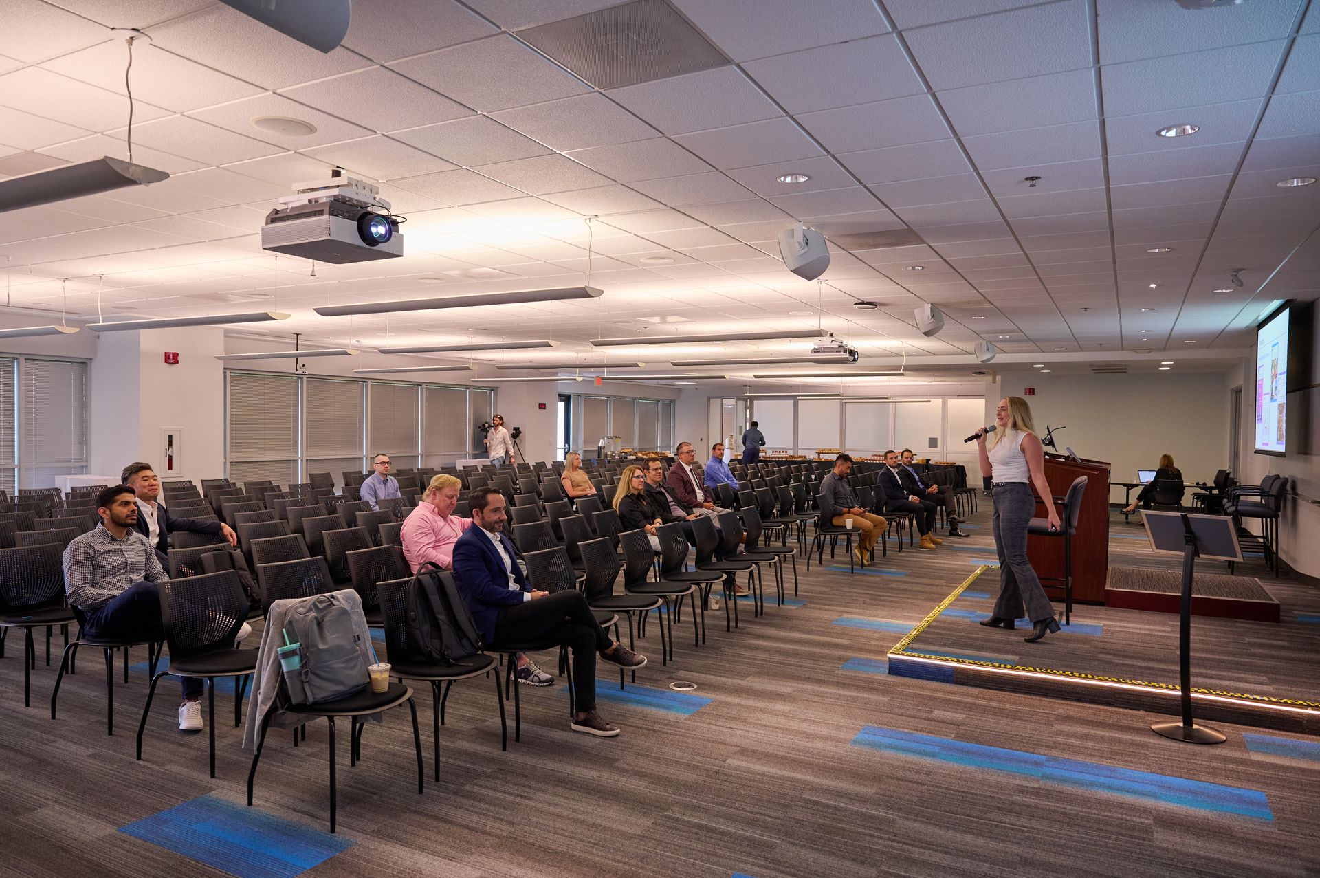 A large room filled with people sitting in chairs and a woman standing at a podium giving a presentation.