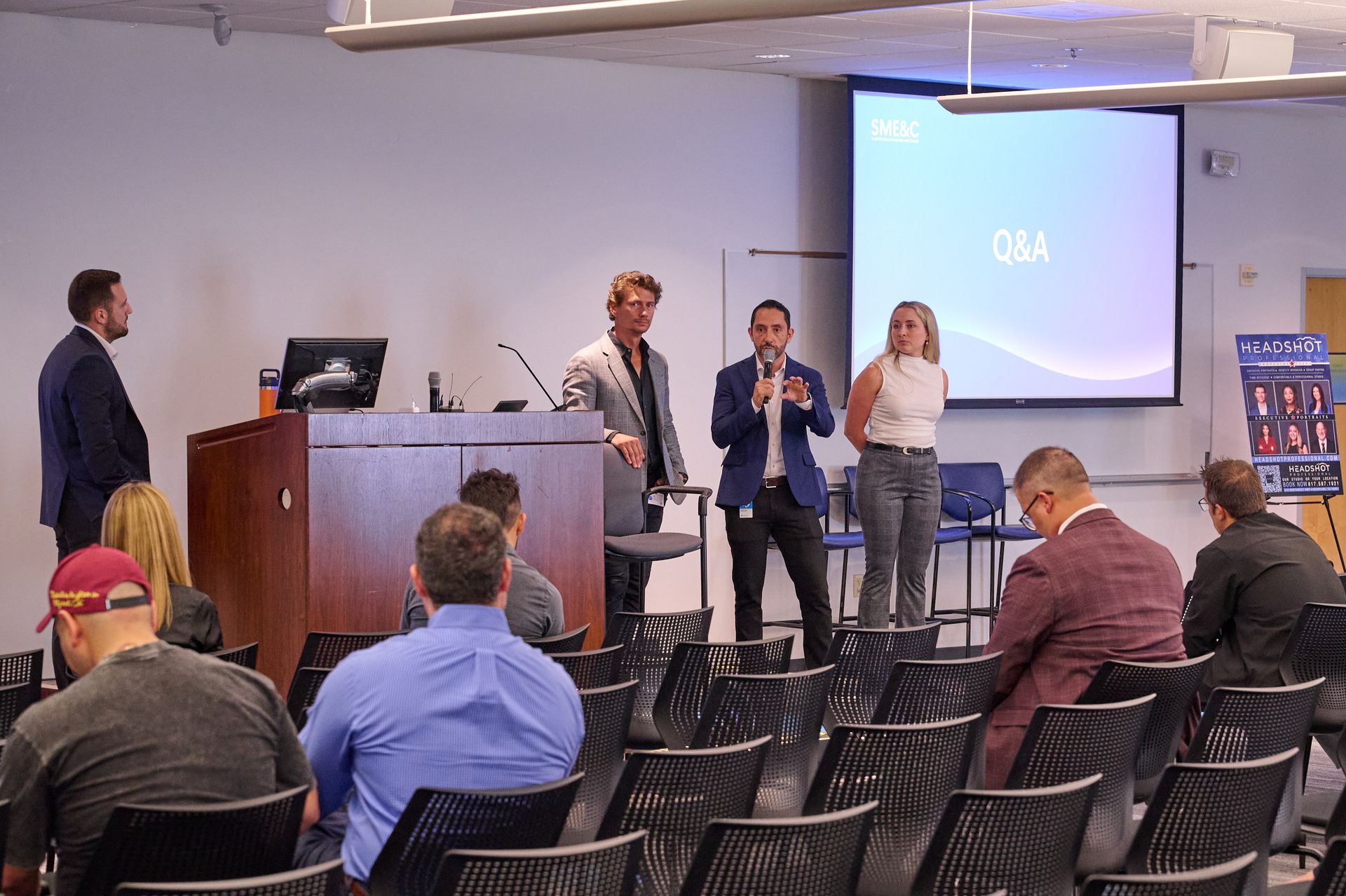 A group of people are sitting in chairs in front of a projector screen in a conference room.