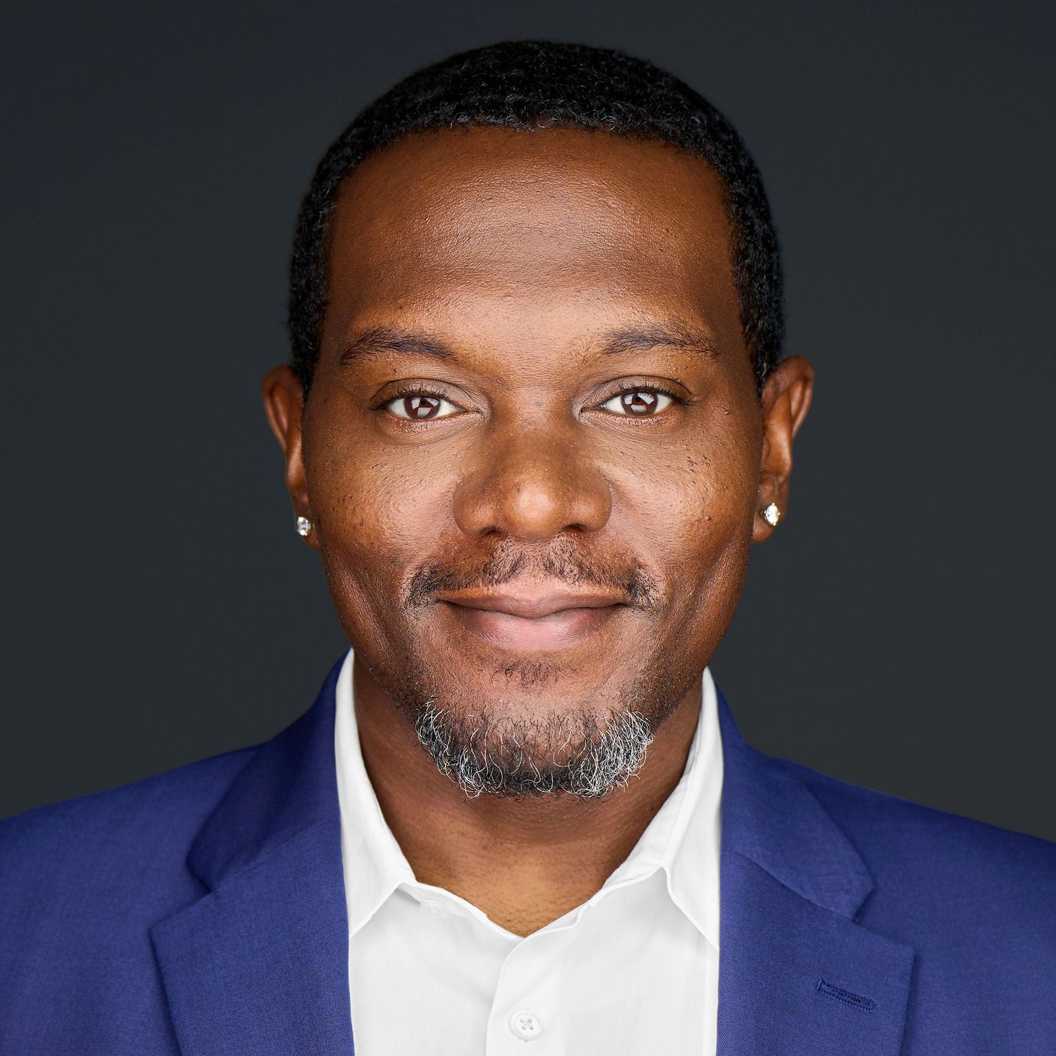 Studio headshot of a person in a blue blazer and white shirt, smiling against a dark background