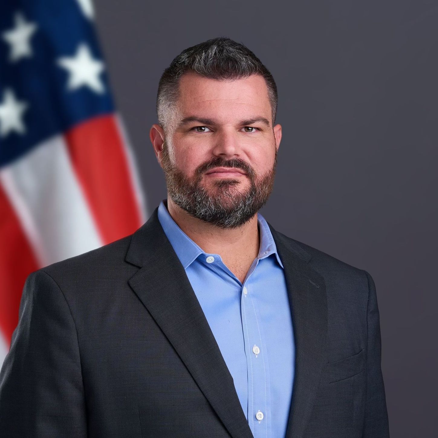 Formal portrait of a man in a blue shirt and dark blazer with a blurred U.S. flag background