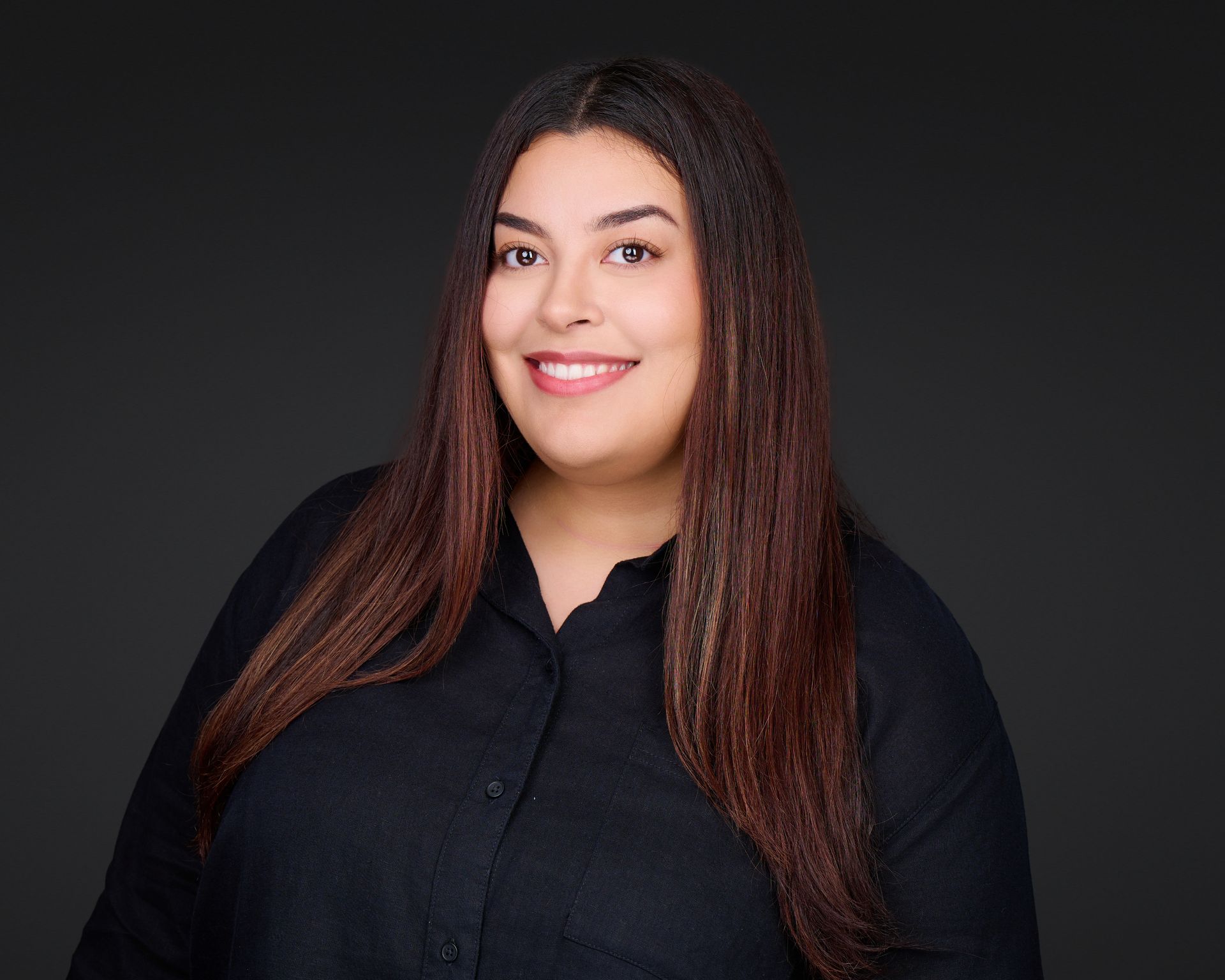 Studio portrait of a smiling person with long dark hair, wearing a black top against a gray background