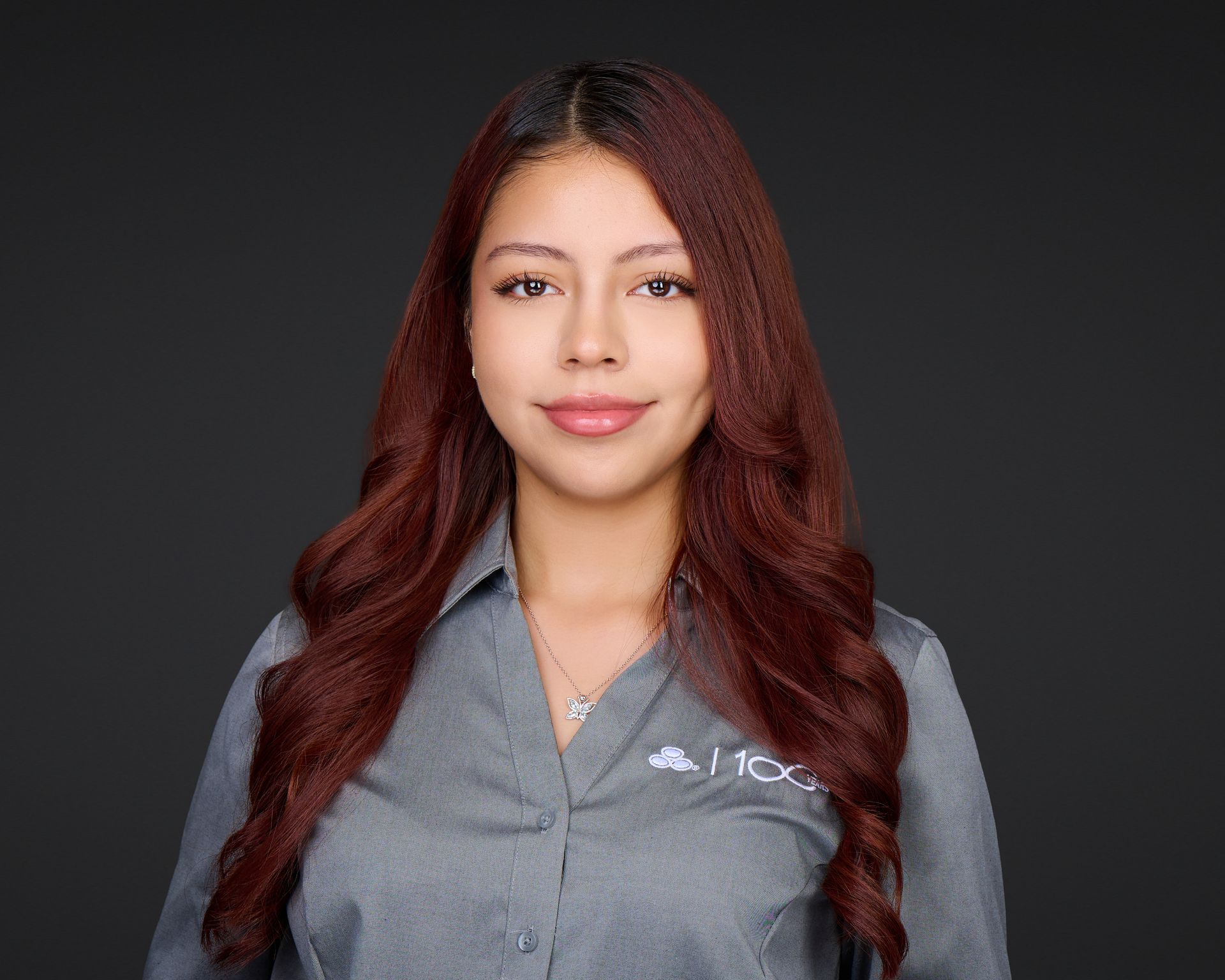 Studio portrait of a woman with long red hair in a gray button-up shirt, smiling against a dark background