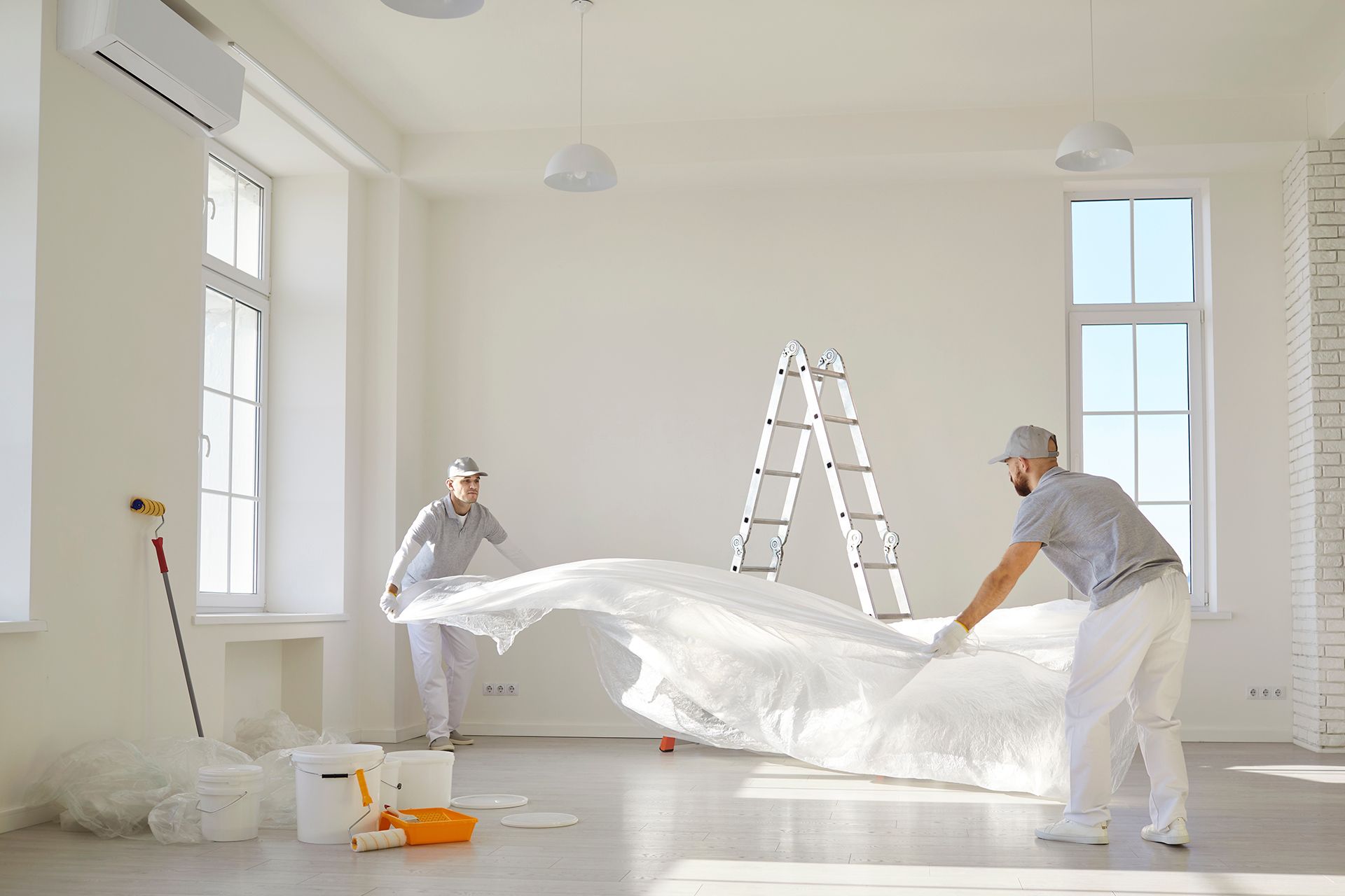Two workers in white coveralls unfold a large plastic sheet in a bright empty room with ladders and painting supplies.