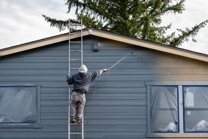 Person on a ladder painting the blue siding of a house under the roof peak