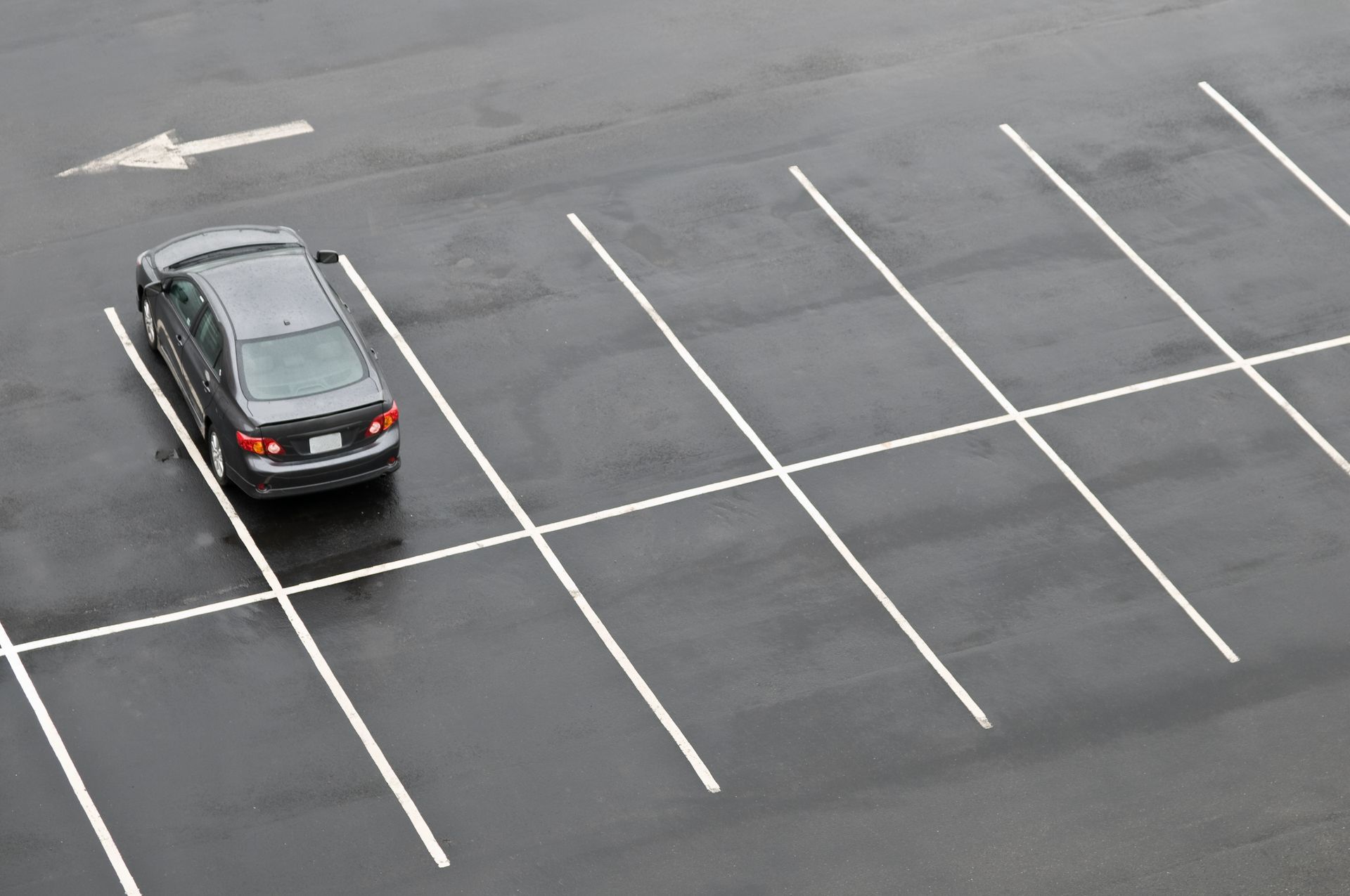A dark car parked in the first spot of a nearly empty, wet asphalt parking lot with white painted lines and an arrow. A dark car parked in the first spot of a nearly empty, wet asphalt parking lot with white painted lines and an arrow.