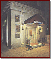 Entrance of a building at night with a lit doorway, awning, and small sign beside the door