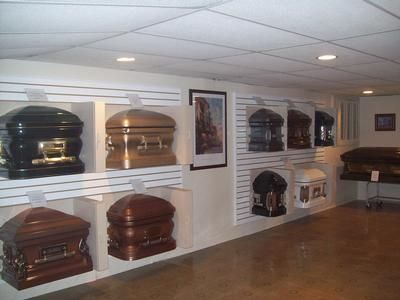 Coffins displayed in a showroom with white walls, wood floor, and framed artwork.