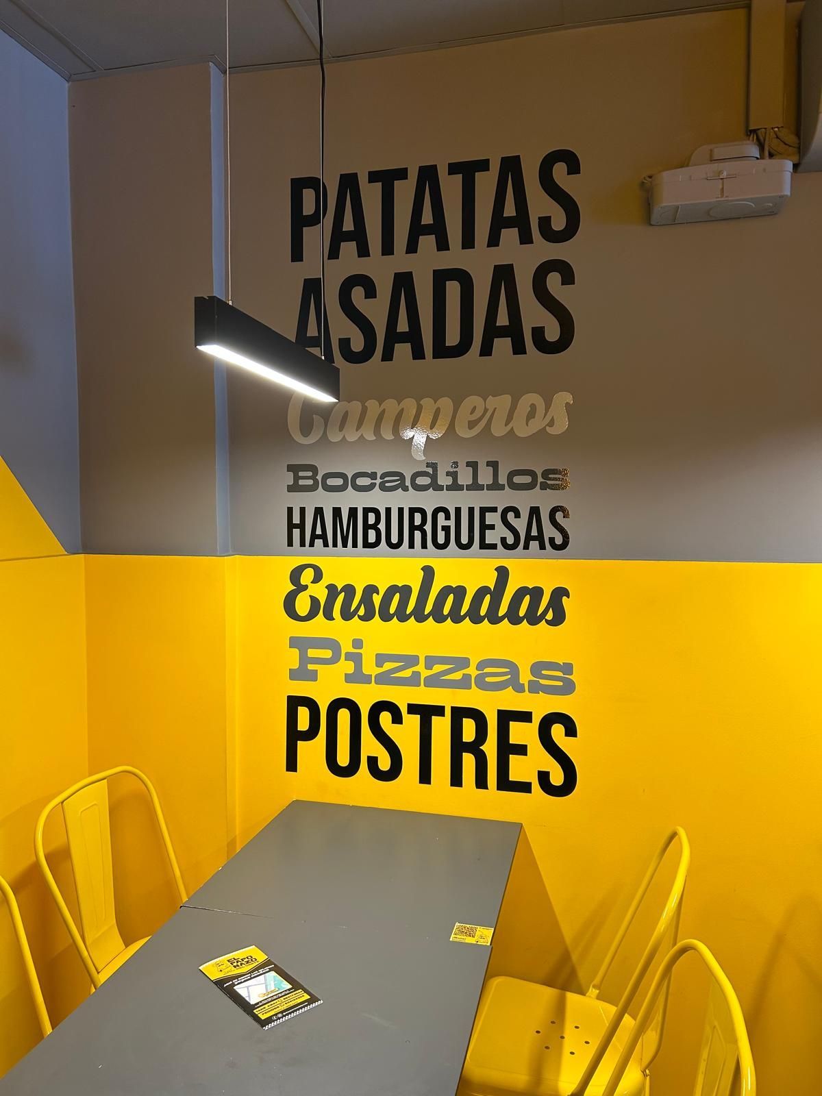 Interior del restaurante con paredes y sillas amarillas, mesa y pizarra con el menú que incluye patatas asadas, hamburguesas, pizzas y postres.