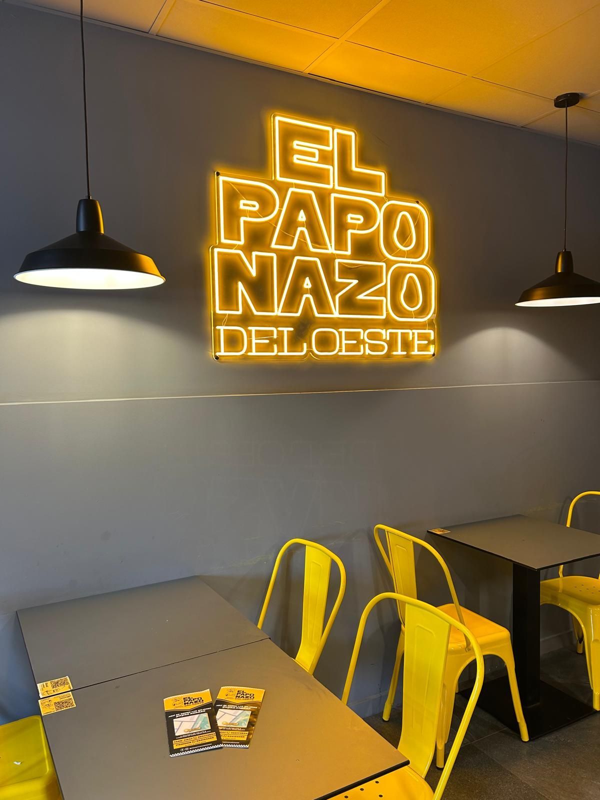 Acogedor rincón de restaurante con un letrero de neón amarillo brillante, lámparas colgantes negras, paredes grises y sillas amarillas.
