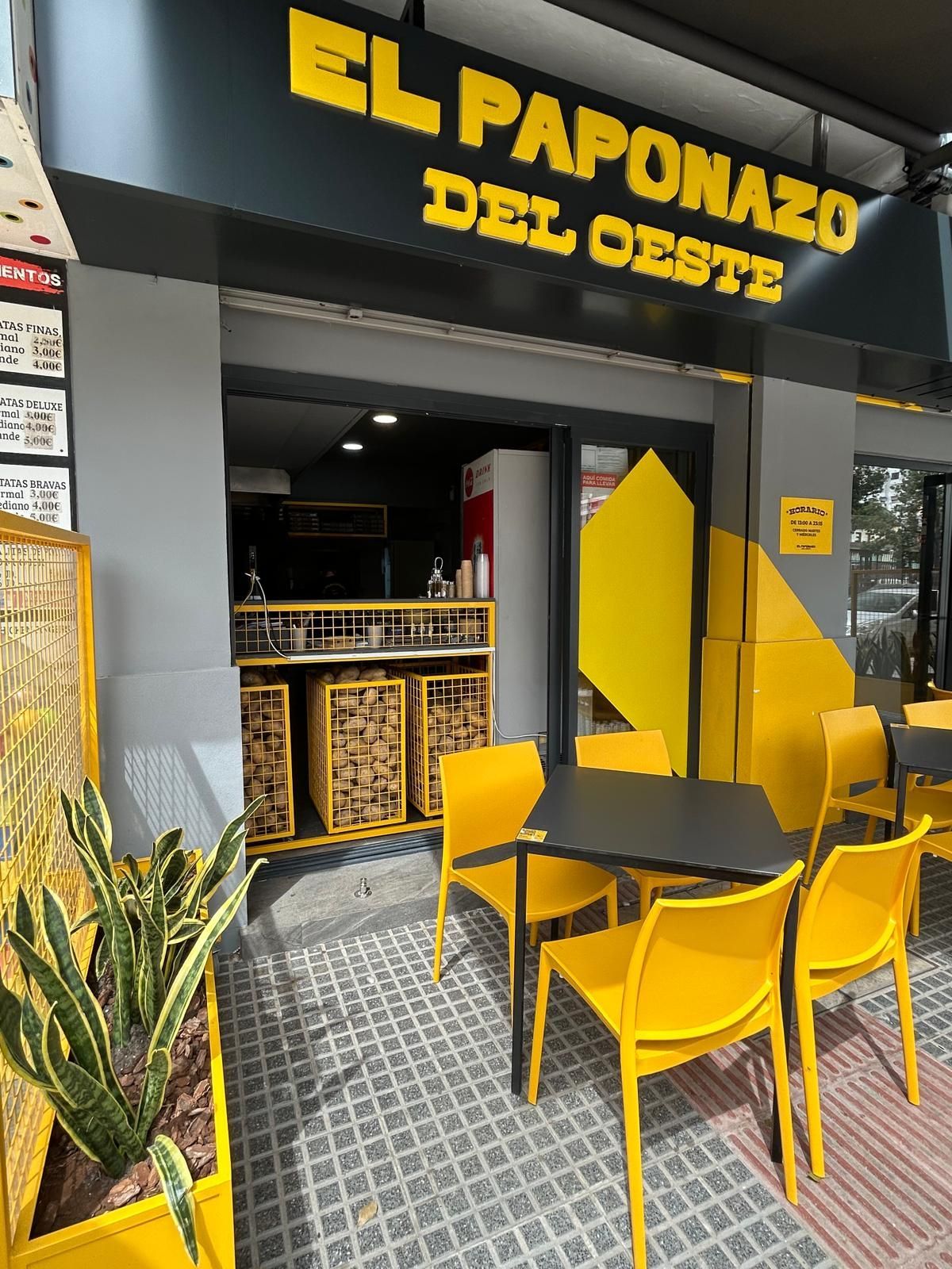 Entrada de un pequeño restaurante con sillas y mesas de color amarillo brillante, y un letrero negro y amarillo que dice “El Paponazo del Oeste”.