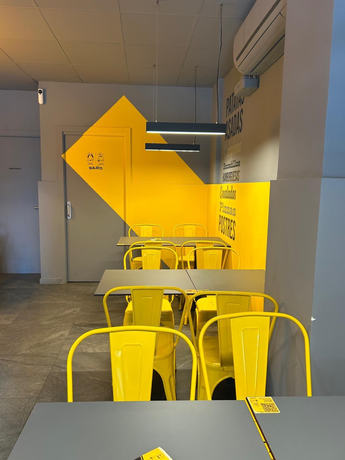 Cafetería pintada de amarillo con sillas y mesas de metal en una estrecha zona de estar interior.
