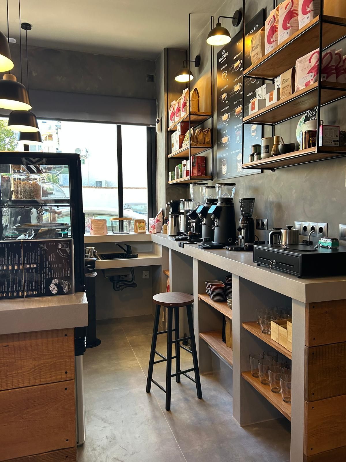 Interior acogedor de una cafetería con máquina de café, estantes con pasteles y otros productos, un taburete junto a la barra y asientos junto a la ventana.