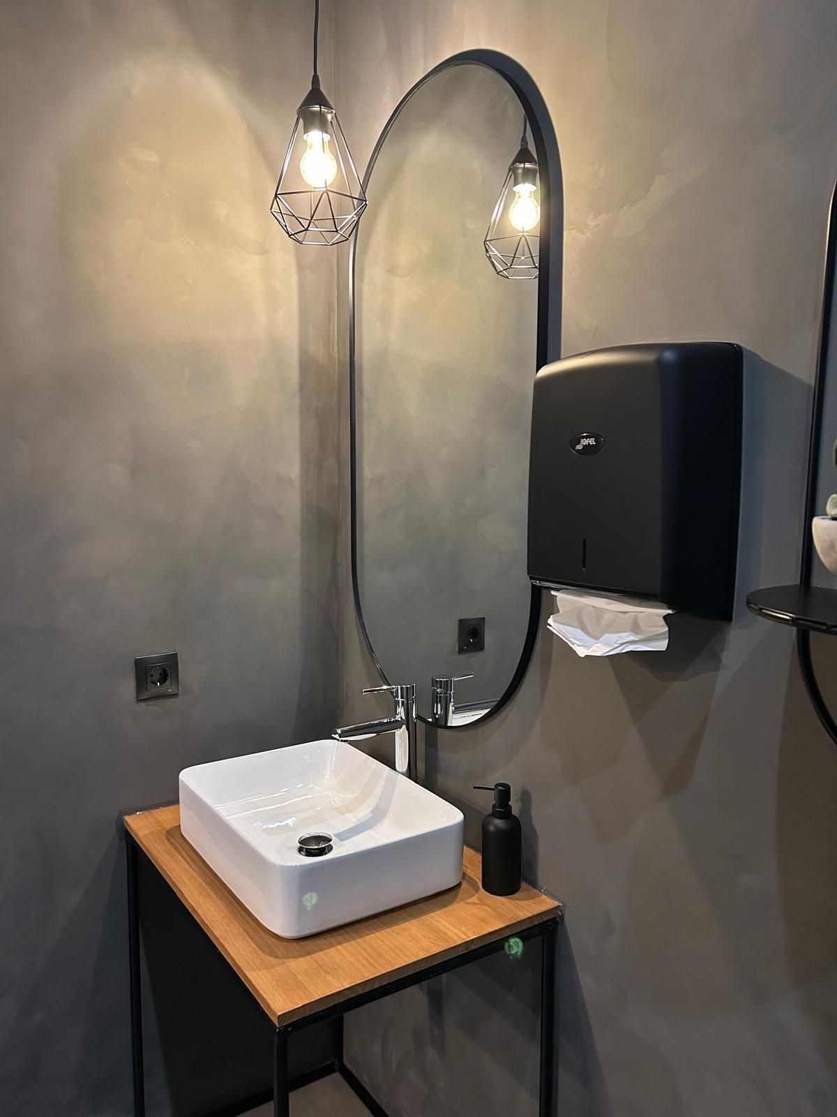 Tocador de baño moderno con lavabo cuadrado, espejo ovalado, apliques de pared y dispensador de toallas de papel negras.