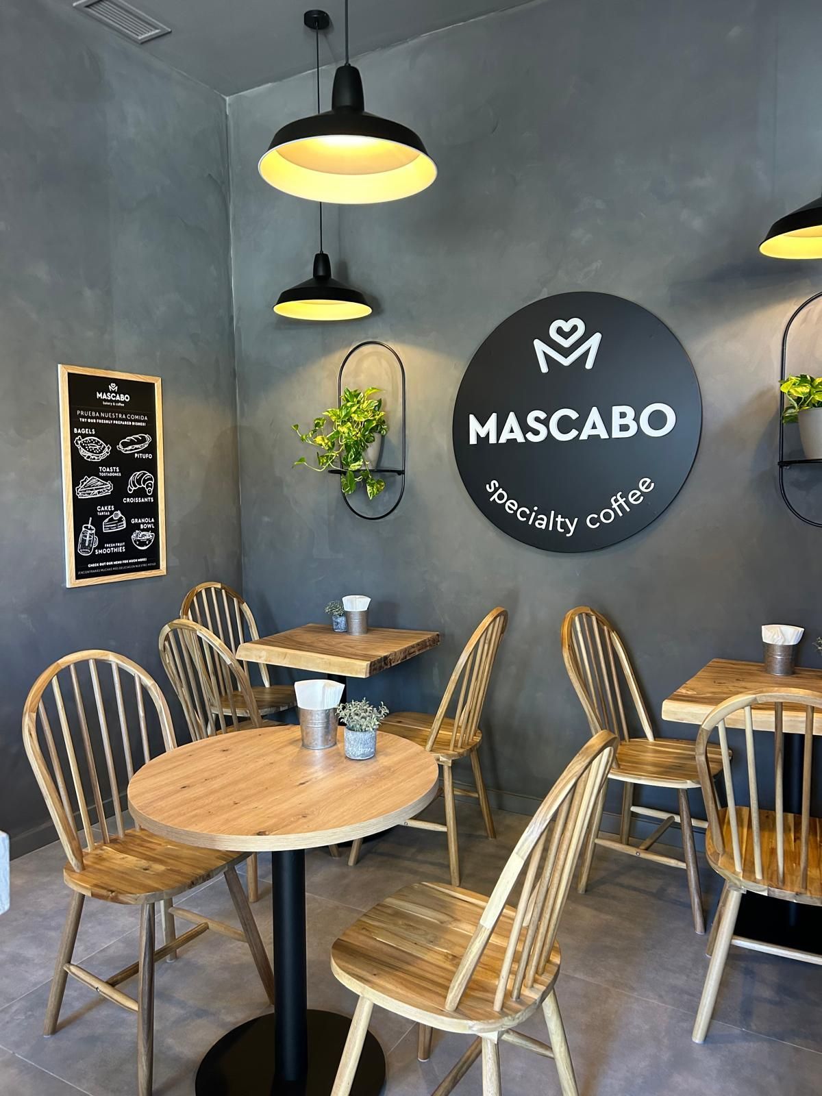 Interior acogedor de una cafetería con mesas redondas de madera, sillas de mimbre y un letrero negro en la pared que dice