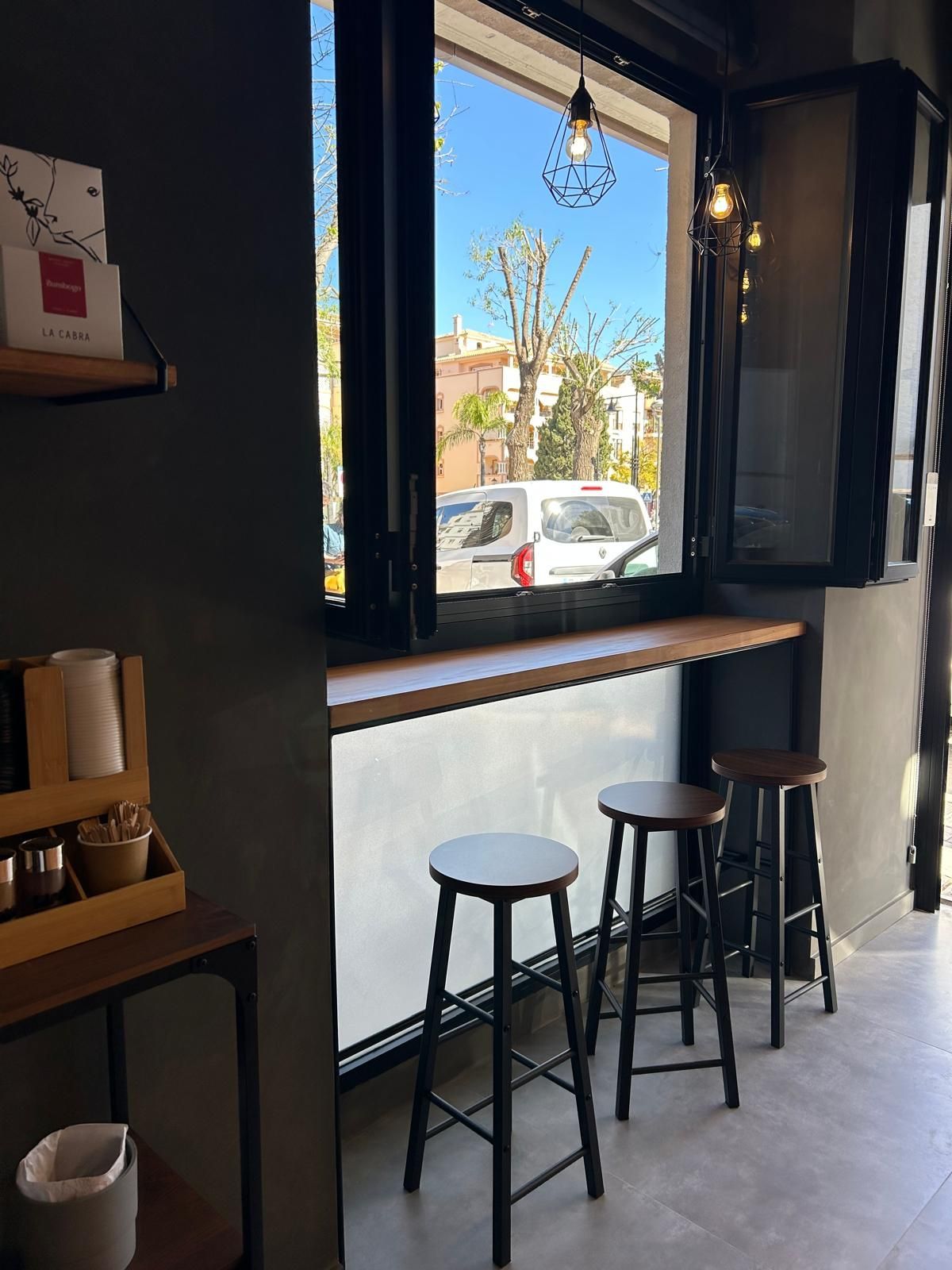 Ventana acogedora de cafetería con tres taburetes, persianas abiertas y vista a la calle.