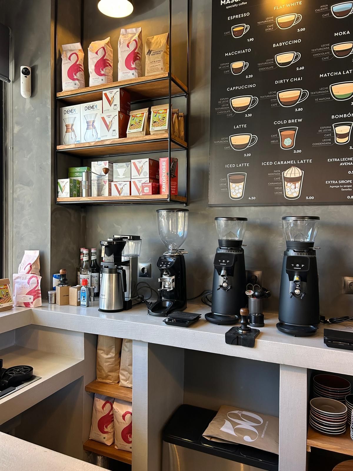 Mostrador de cafetería con máquinas de espresso, tazas y estantes con productos de café junto a una pizarra con el menú.