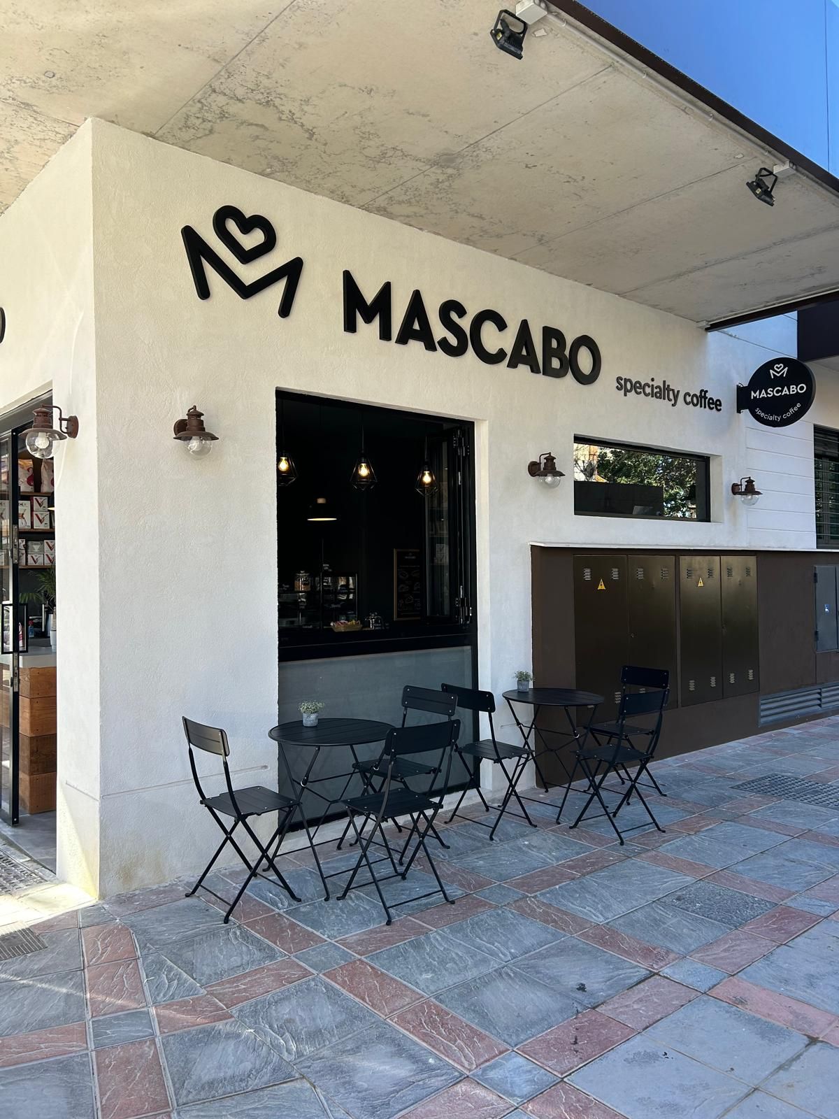 Exterior de la cafetería Mascabo con letrero negro, mesa y sillas al aire libre y zona de asientos en la acera con baldosas.