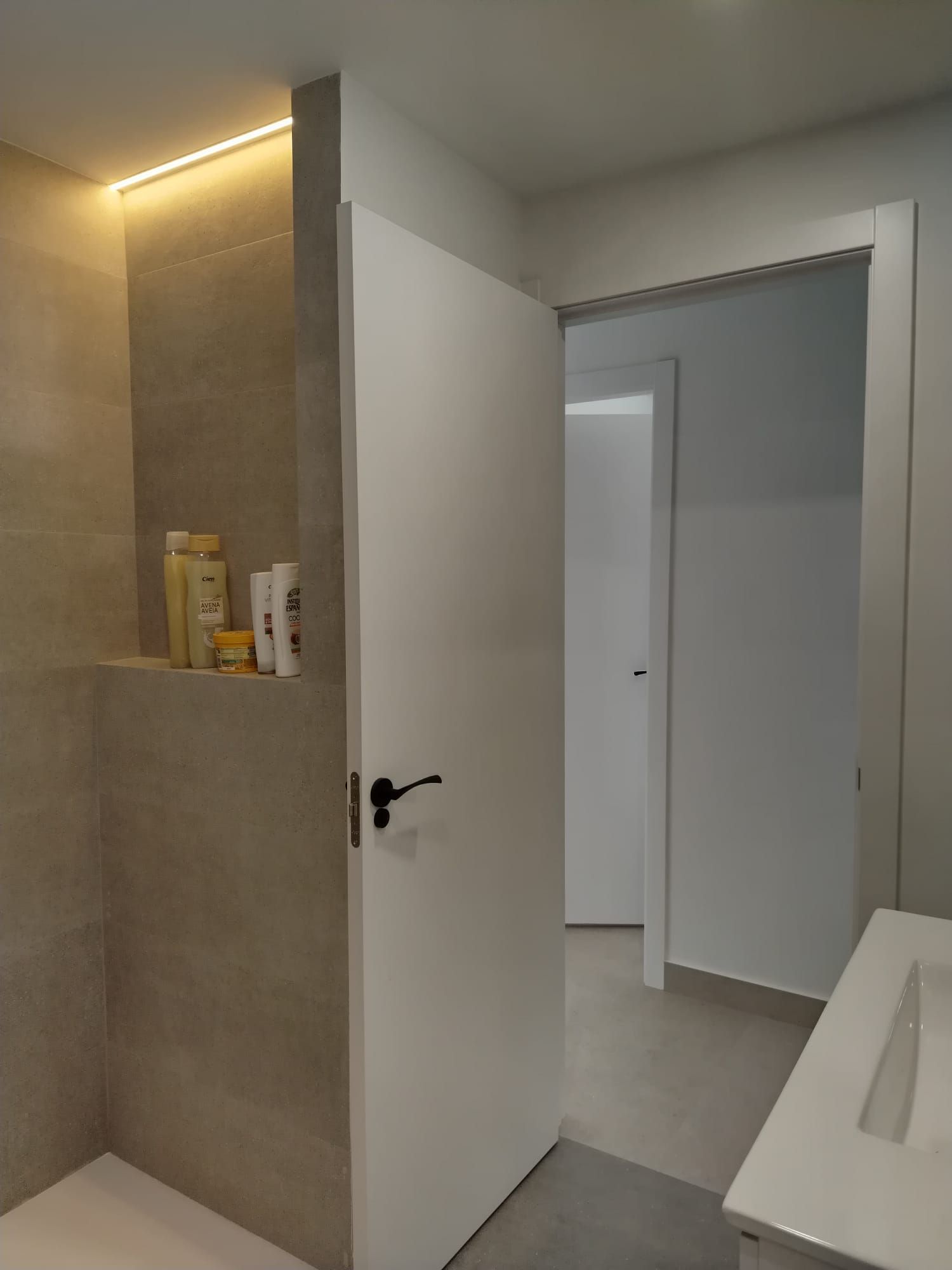 Puerta del baño abierta que deja ver una pared de azulejos grises, un estante para artículos de tocador y un lavabo en una habitación luminosa.