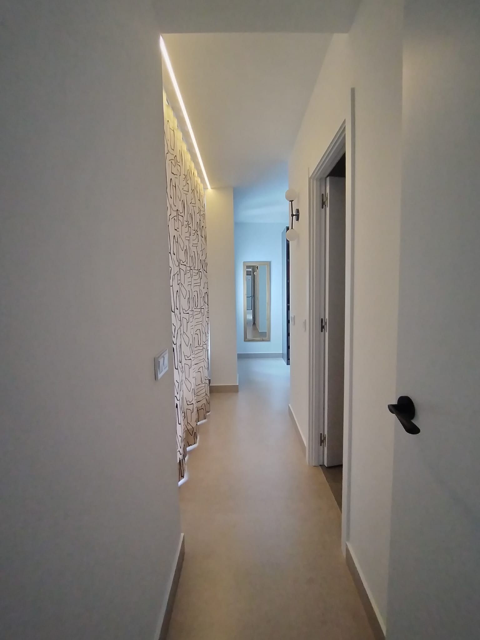 Un estrecho pasillo blanco con puertas y una cortina estampada, que conduce a una habitación luminosa al final.