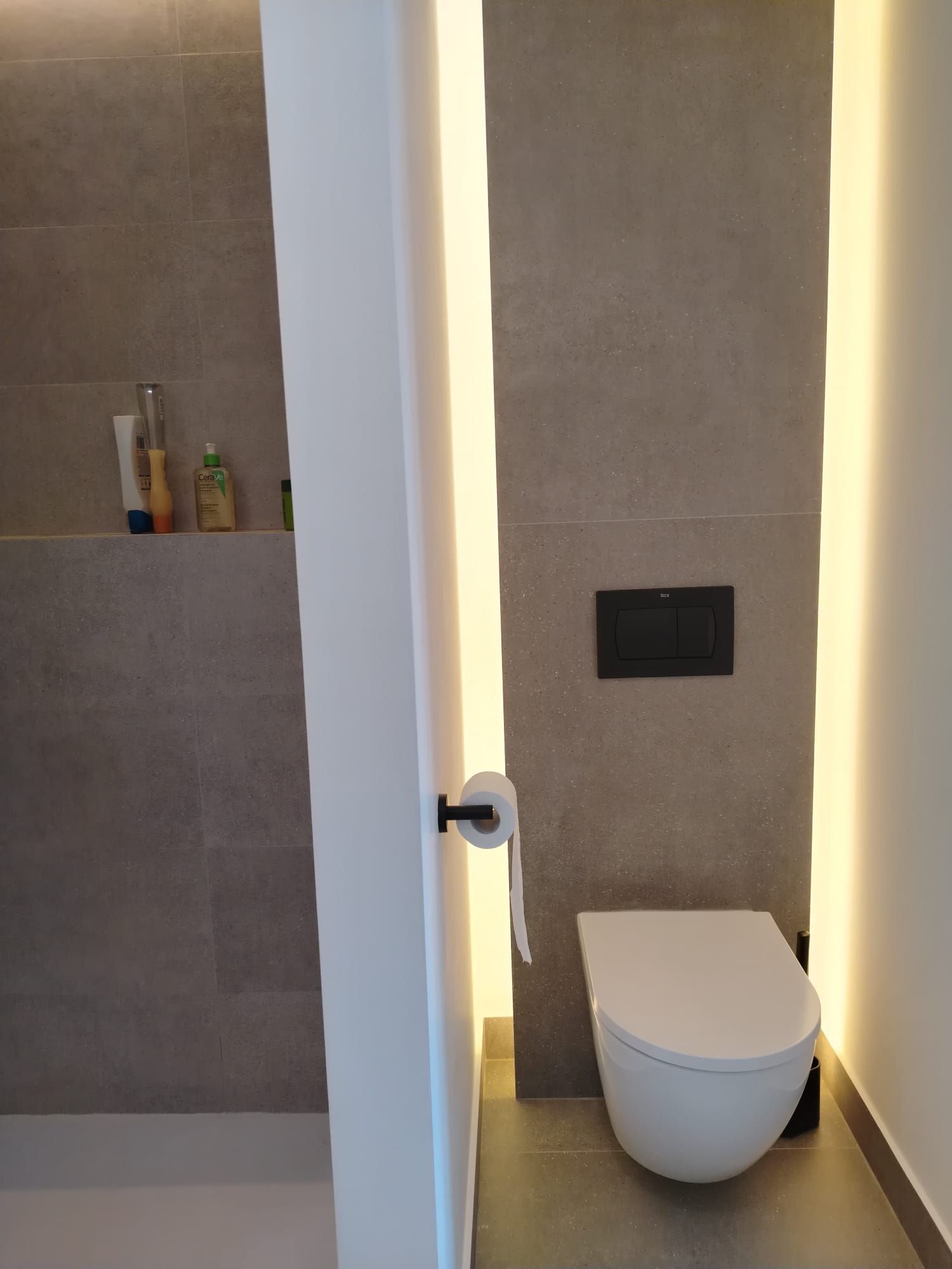 Baño moderno con inodoro, panel de descarga mural y estante de ducha en azulejos grises.