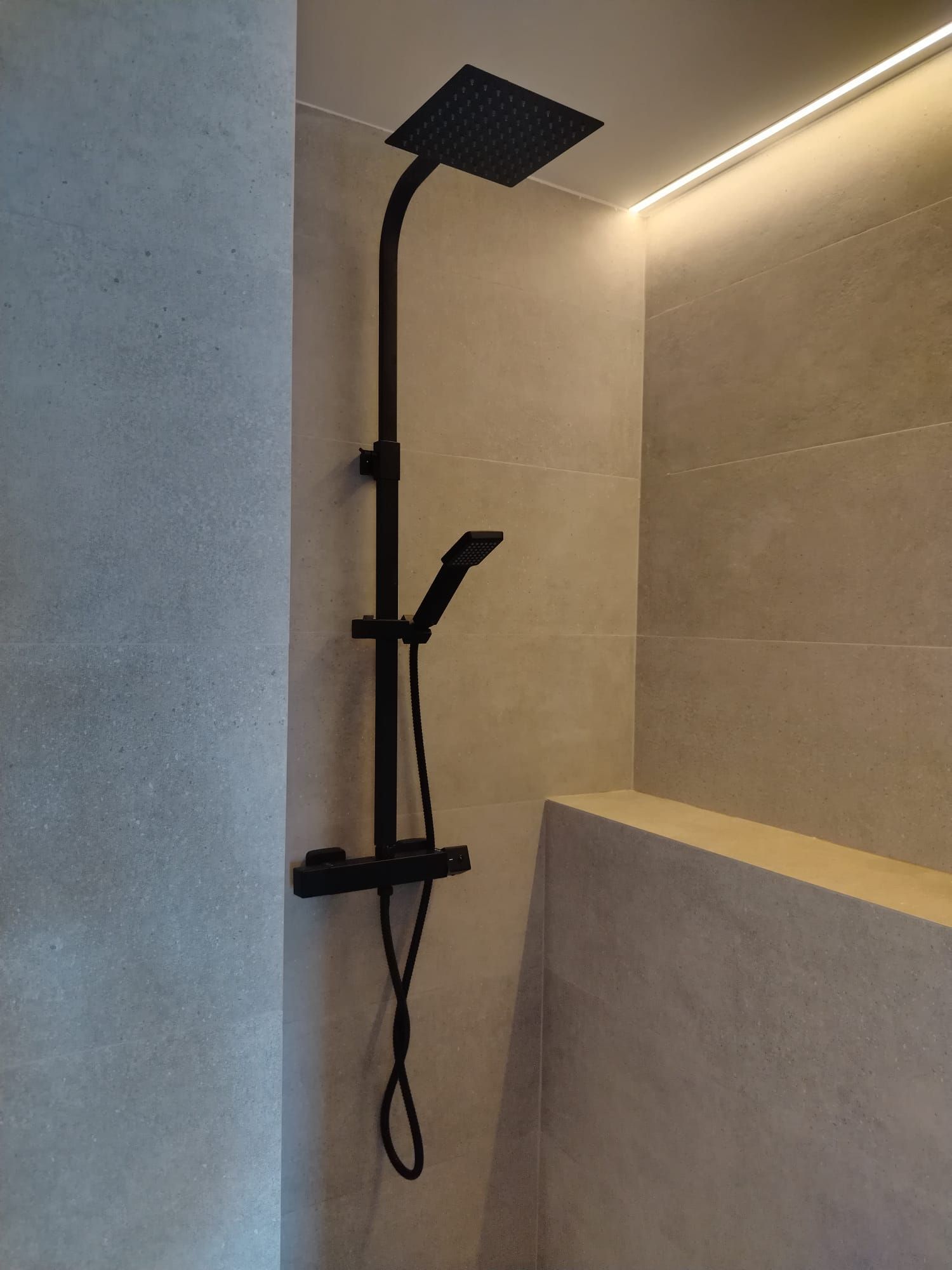 Cabezal de ducha negro y rociador de mano en una cabina de ducha con azulejos beige.
