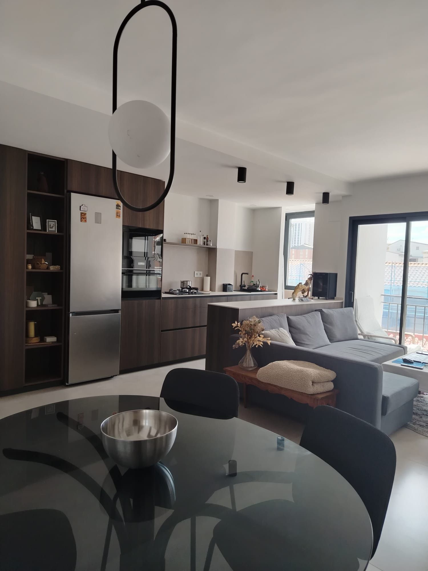 Salón y cocina modernos de planta abierta con sofá gris, mesa de comedor de cristal y grandes ventanales.