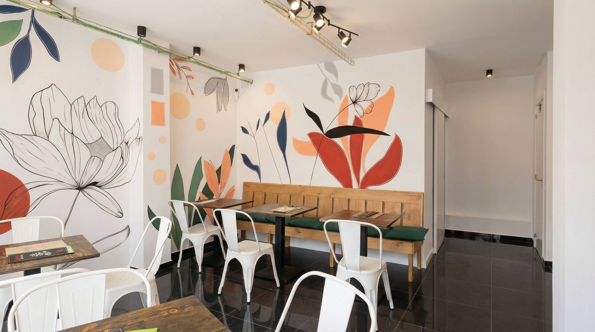 Interior de cafetería moderna con paredes decoradas con murales florales, mesas de madera y sillas blancas.