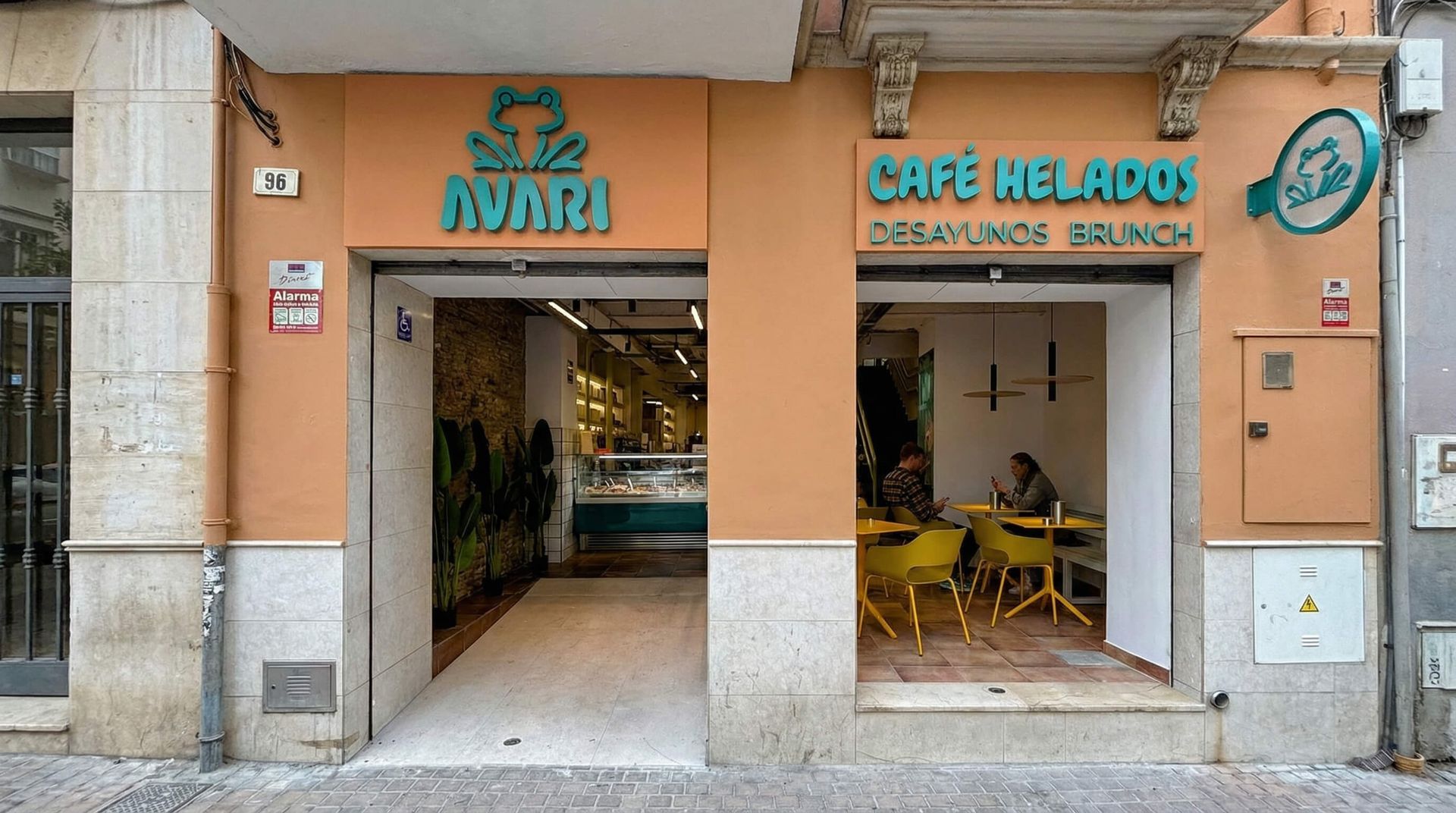 Fachada de cafetería con letrero turquesa, fachada color melocotón y mesas amarillas dentro de una puerta abierta.