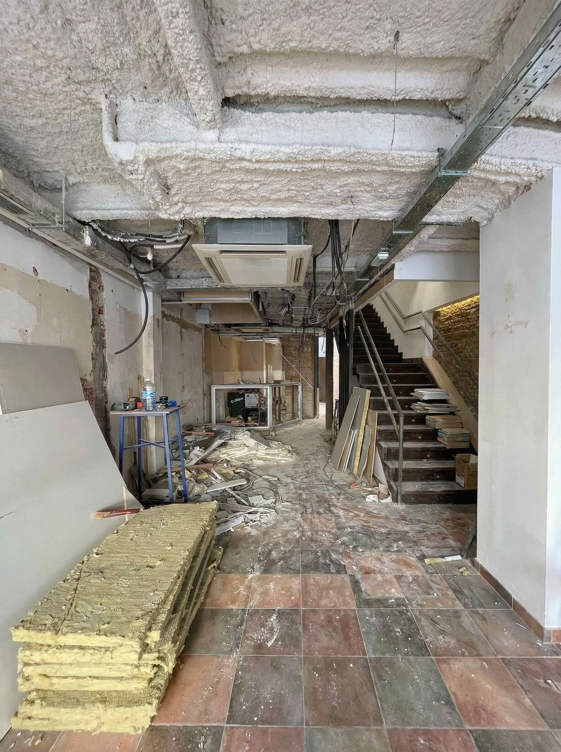 Pasillo interior dañado con escombros, techo al descubierto, escaleras y materiales de construcción.