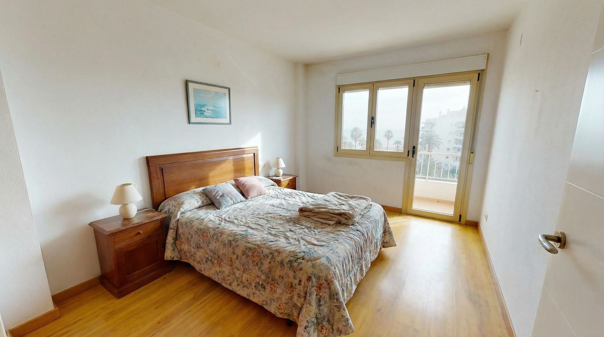 Habitación luminosa con cama de madera, mesita de noche y puertas de cristal que dan a un balcón.