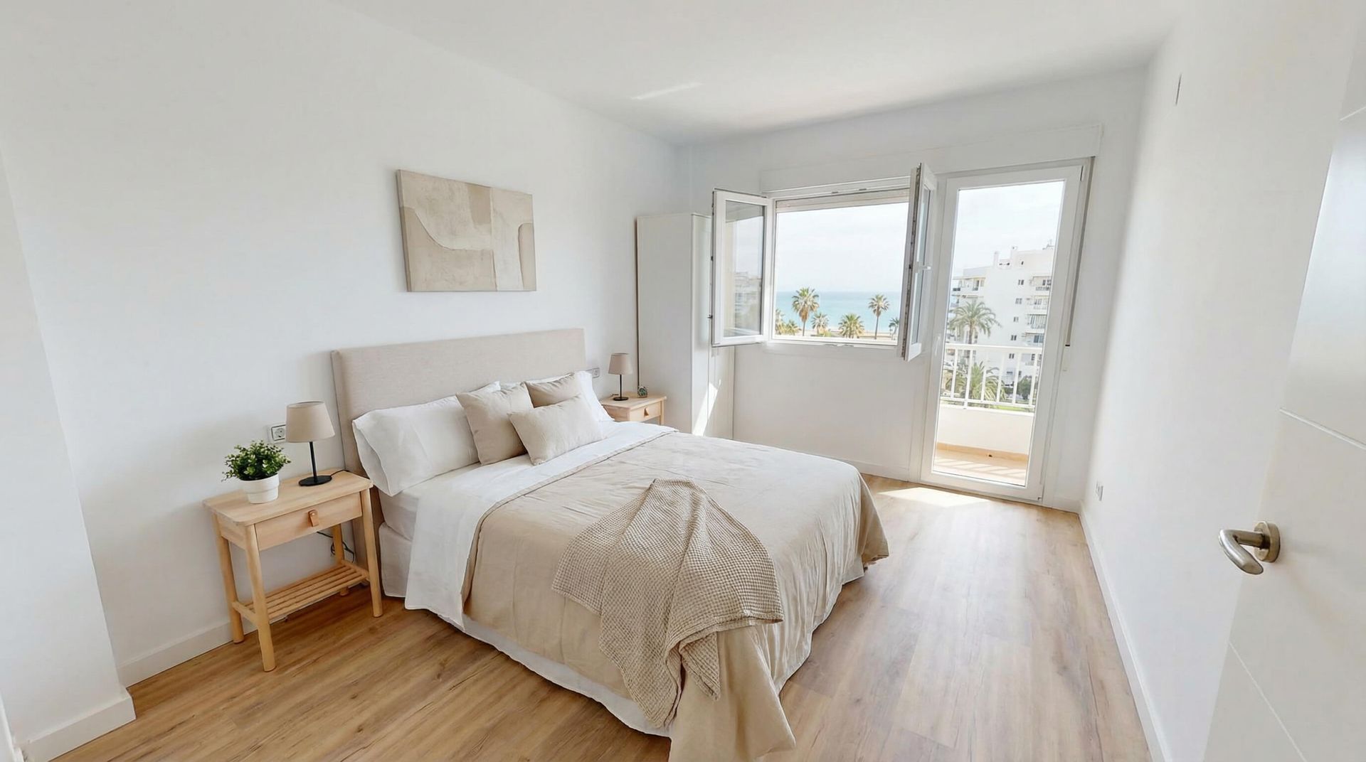Habitación luminosa con ropa de cama blanca, suelo de madera y puertas que dan a un balcón soleado.