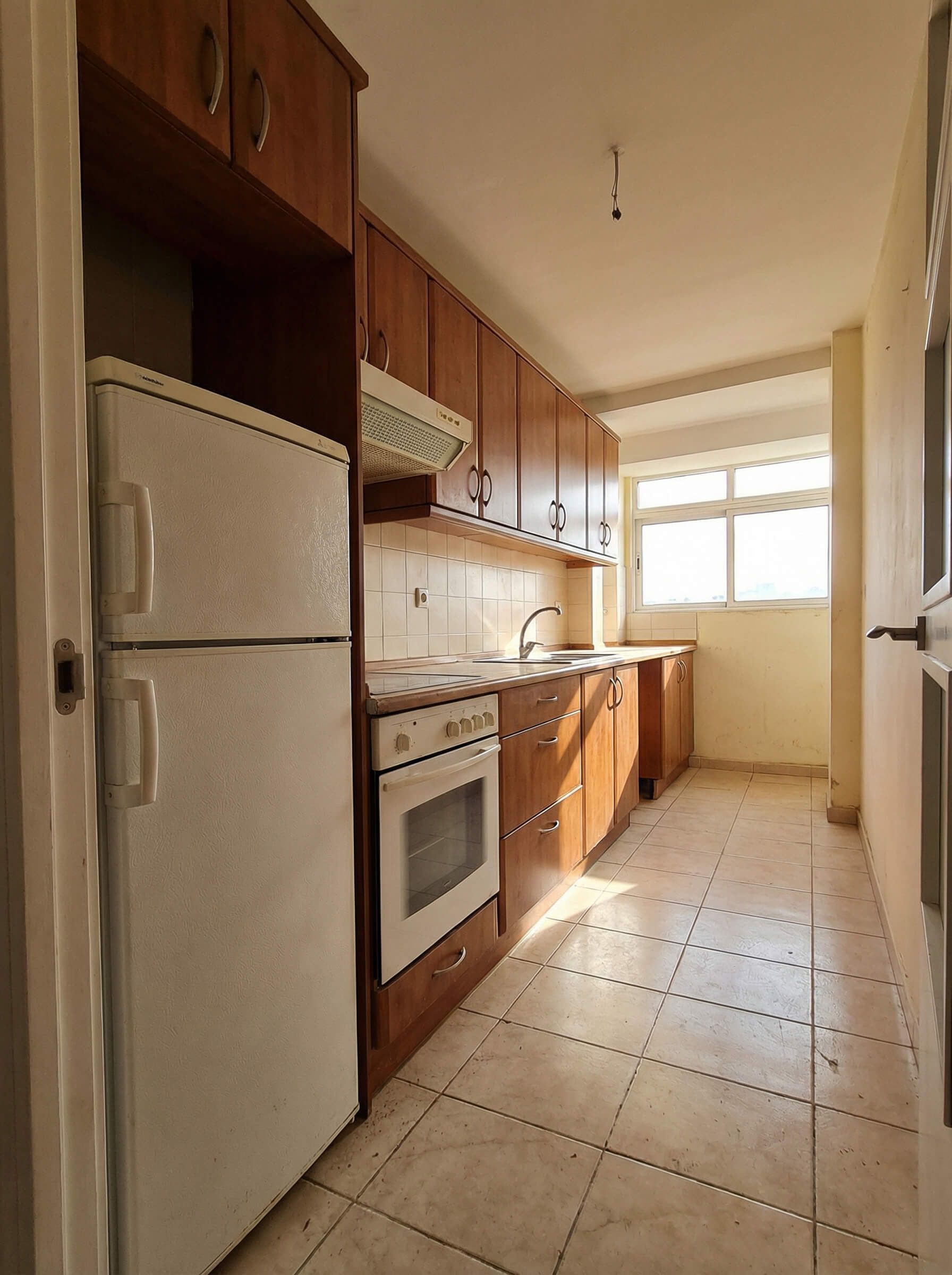 Cocina estrecha con armarios de madera, electrodomésticos blancos, suelo de baldosas beige y una ventana luminosa al final.