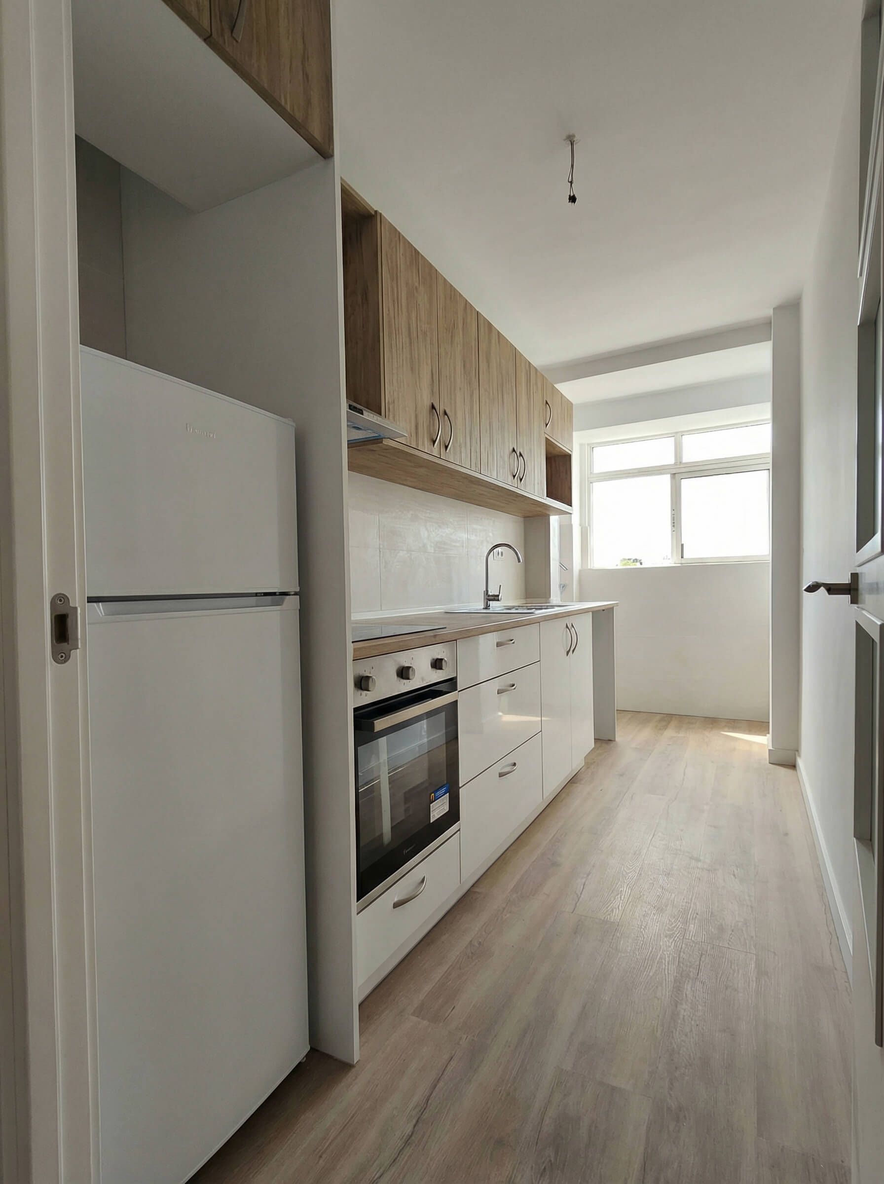 Cocina estrecha y luminosa, tipo galera, con armarios blancos, detalles de madera y una gran ventana al final.