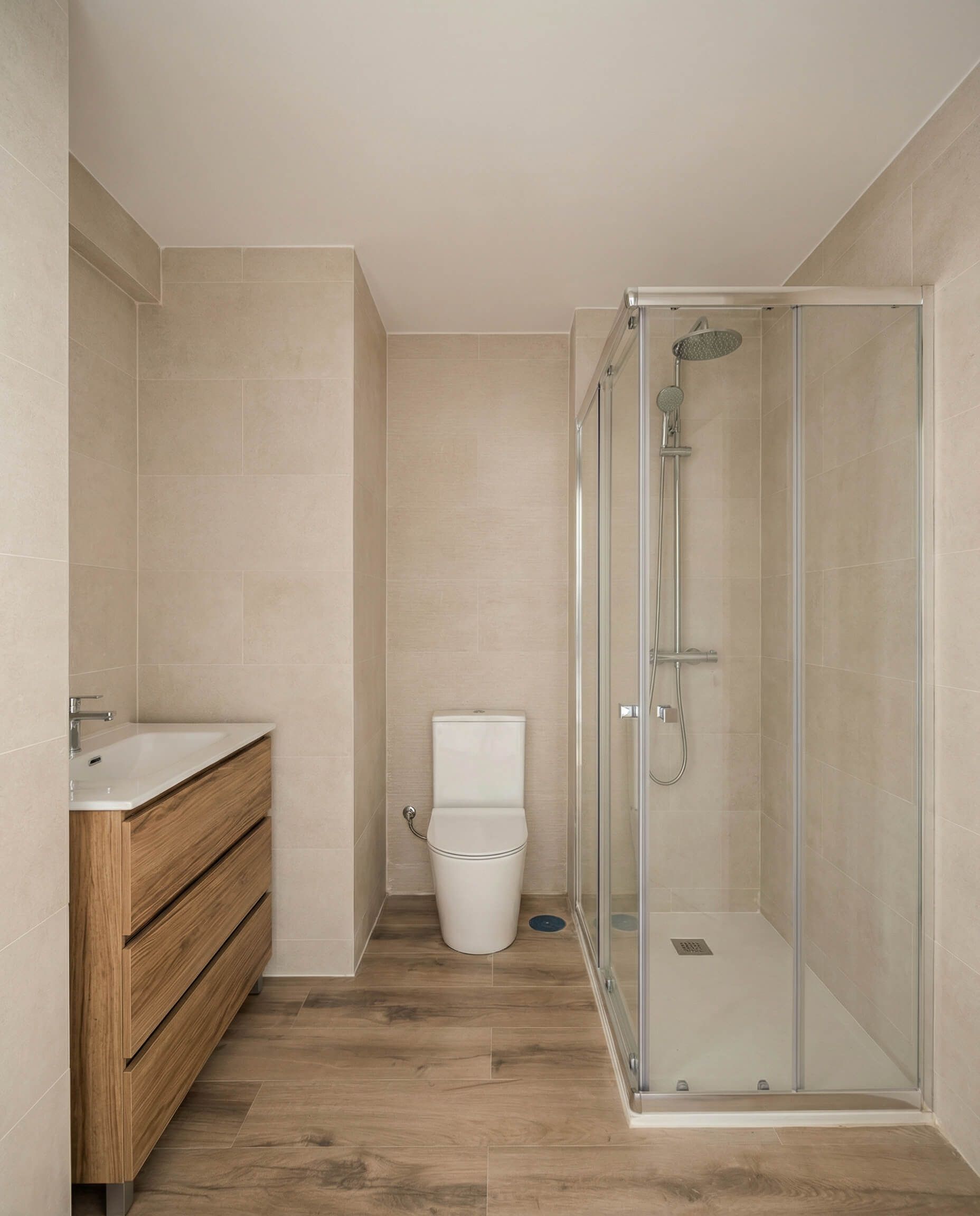 Baño moderno de color beige con lavabo de madera, inodoro y cabina de ducha de cristal.