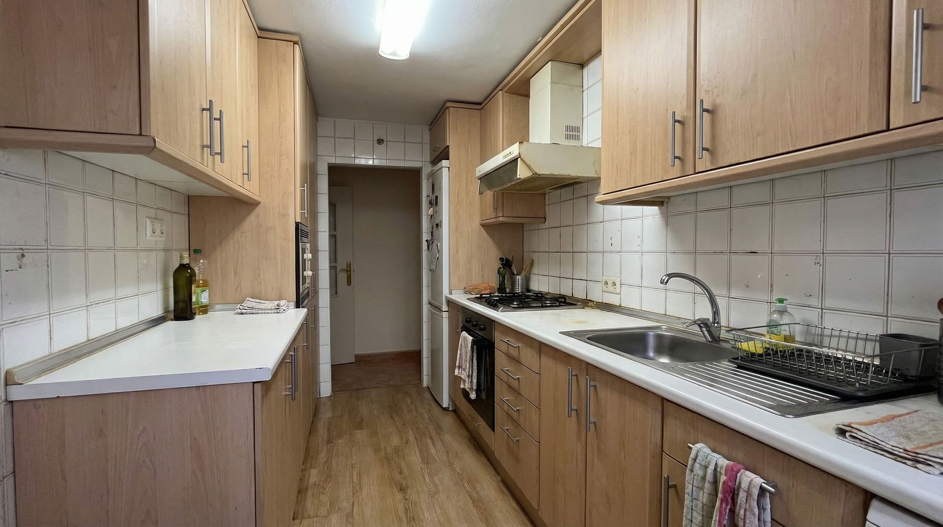 Cocina estrecha tipo galera con gabinetes de madera, encimeras blancas y salpicadero de azulejos, vista hacia un pasillo.
