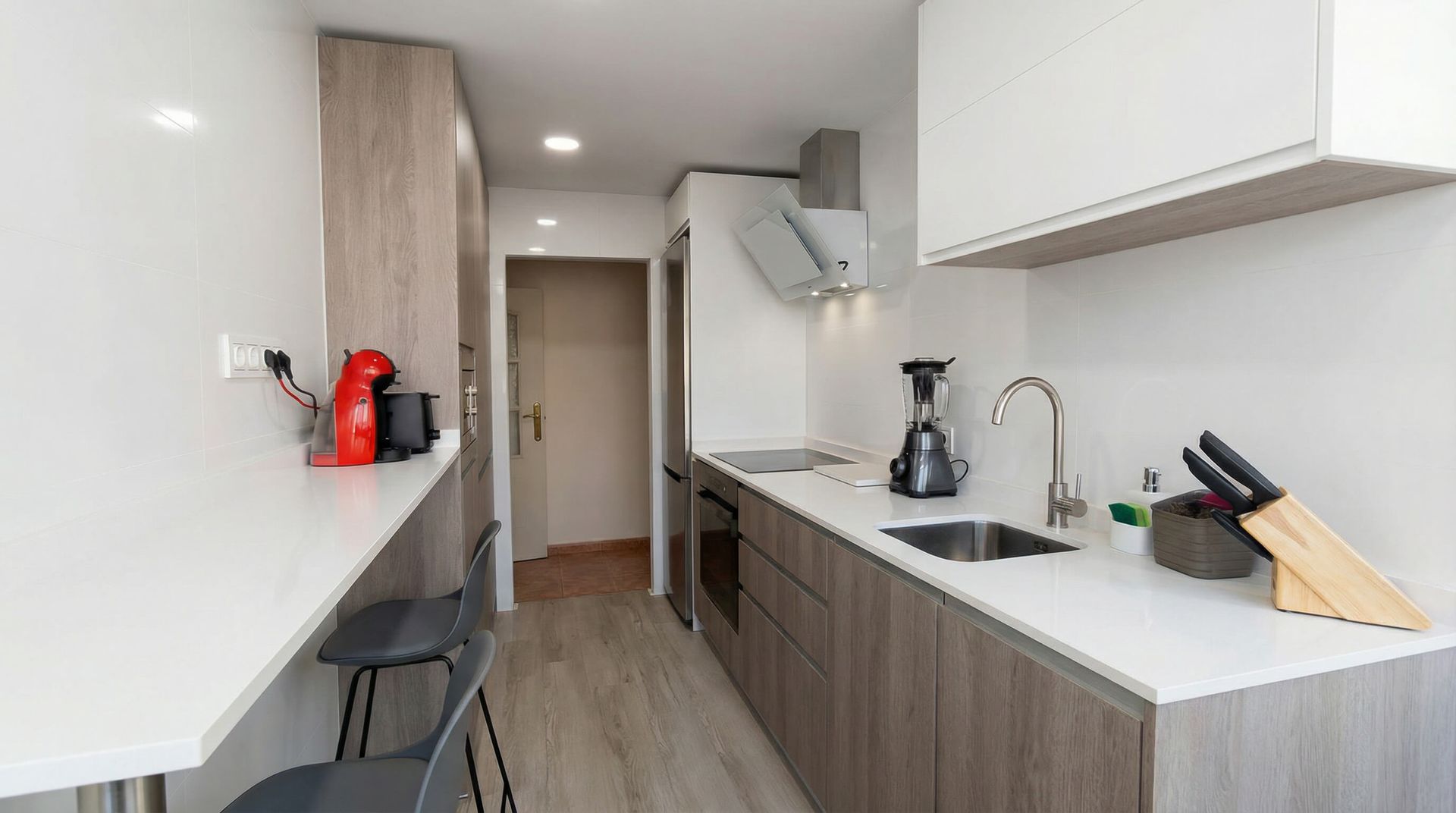 Cocina moderna y estrecha con armarios blancos, suelo de madera, encimera, fogones, fregadero y una pequeña mesa junto a la pared.