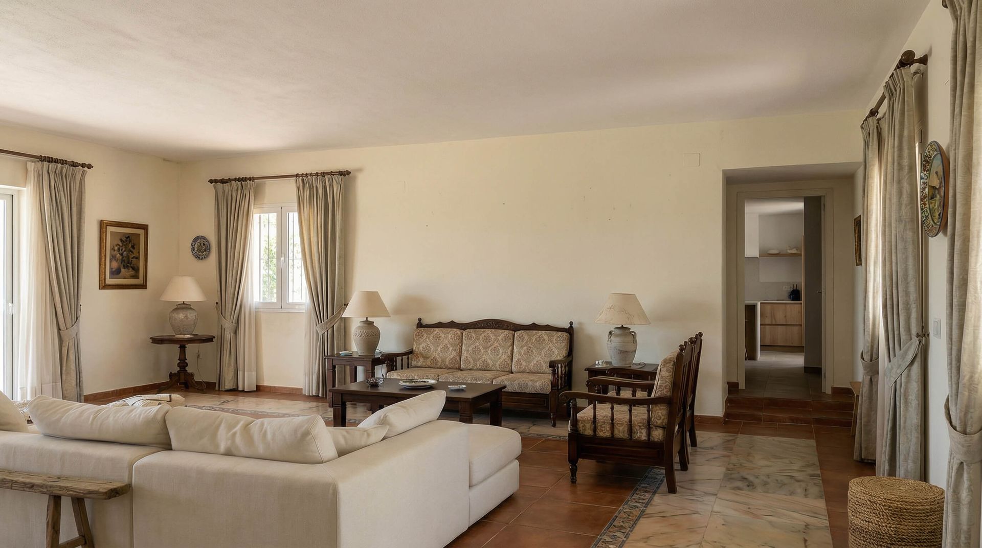 Amplia sala de estar con sofás color crema, sillas estampadas, mesas auxiliares y una puerta abierta que comunica con otra habitación.