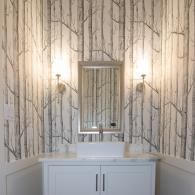 A modern bathroom vanity with a white basin sink, rectangular mirror, and two wall sconces against forest wallpaper.
