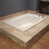 A white whirlpool bathtub set into a beige stone platform, surrounded by matching tiled flooring.